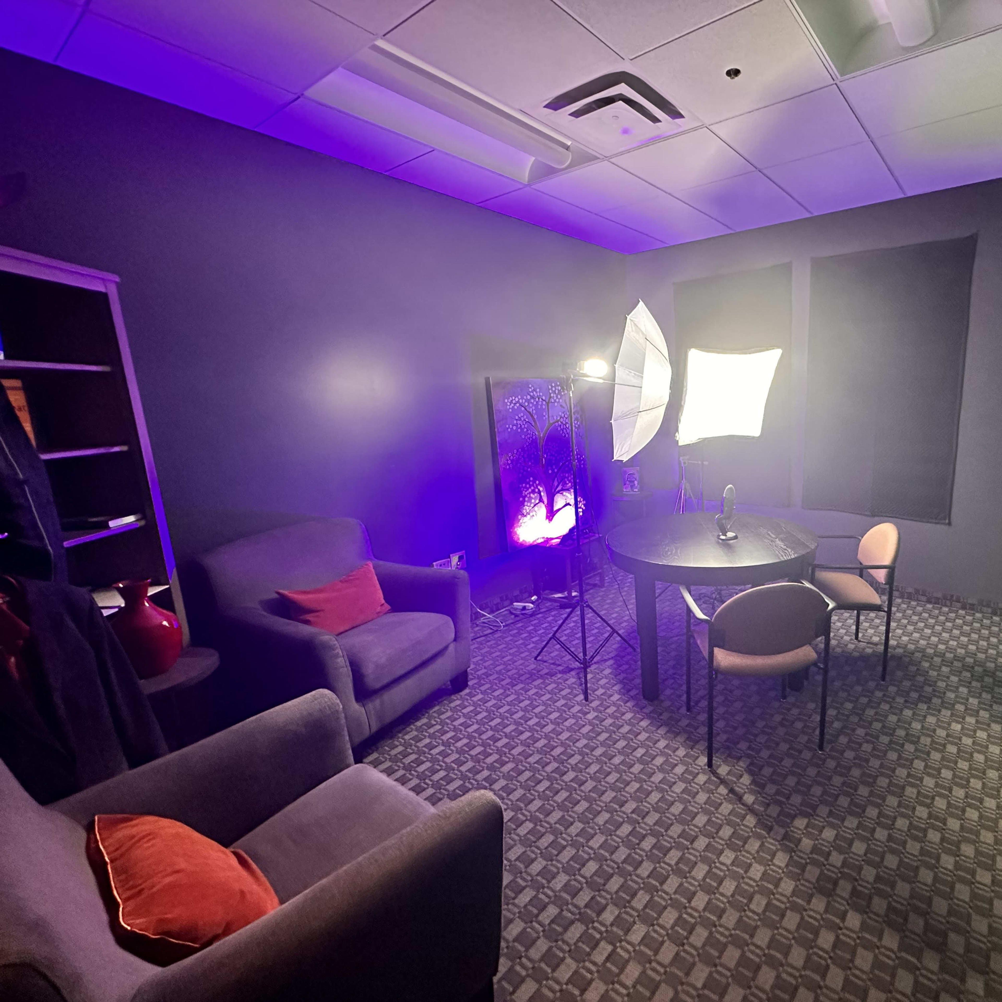 A small studio room with a green screen, a round table, two chairs, and softbox lights positioned for video recording.