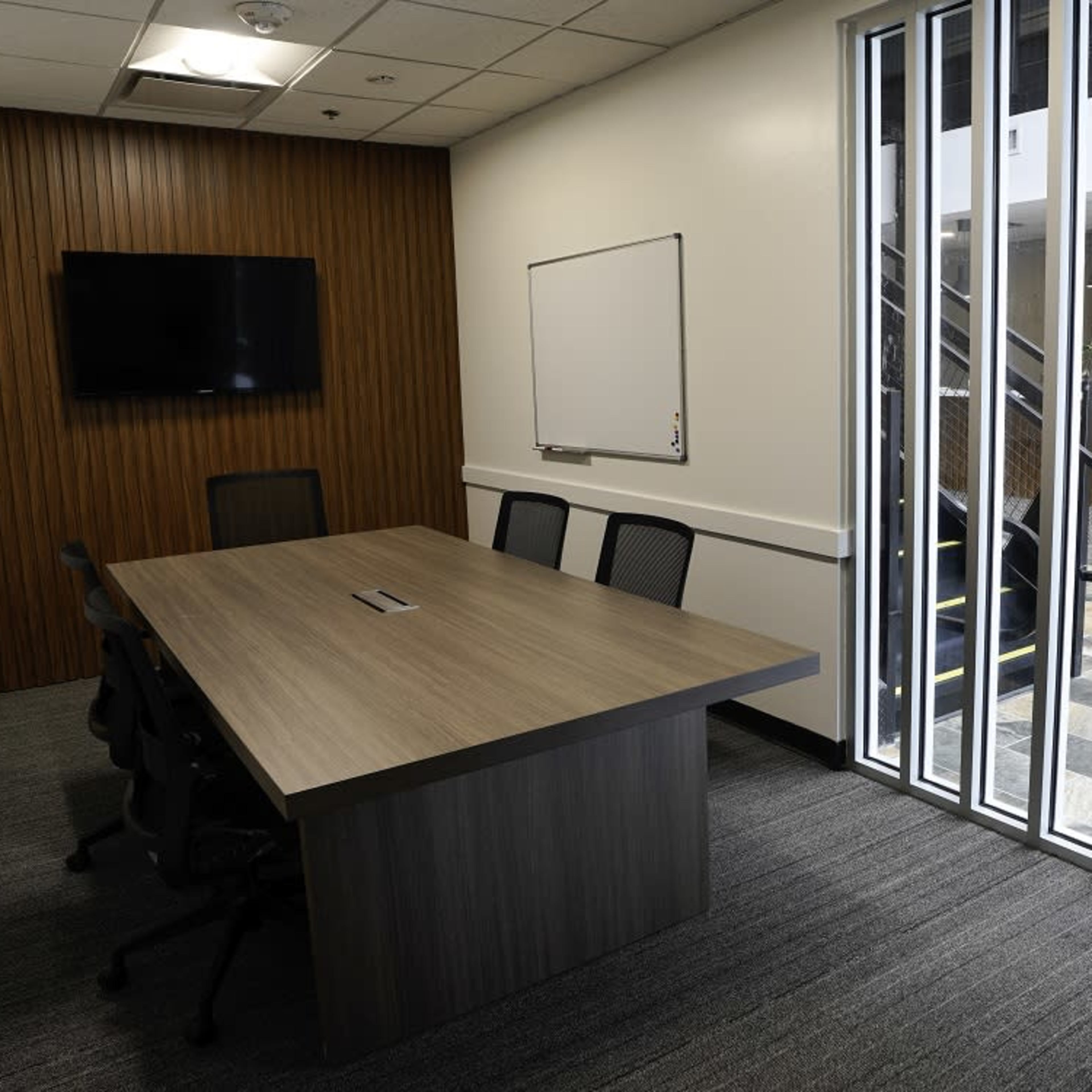 A modern meeting room features a large table surrounded by chairs, a wall-mounted television, and large windows leading to an outdoor area.