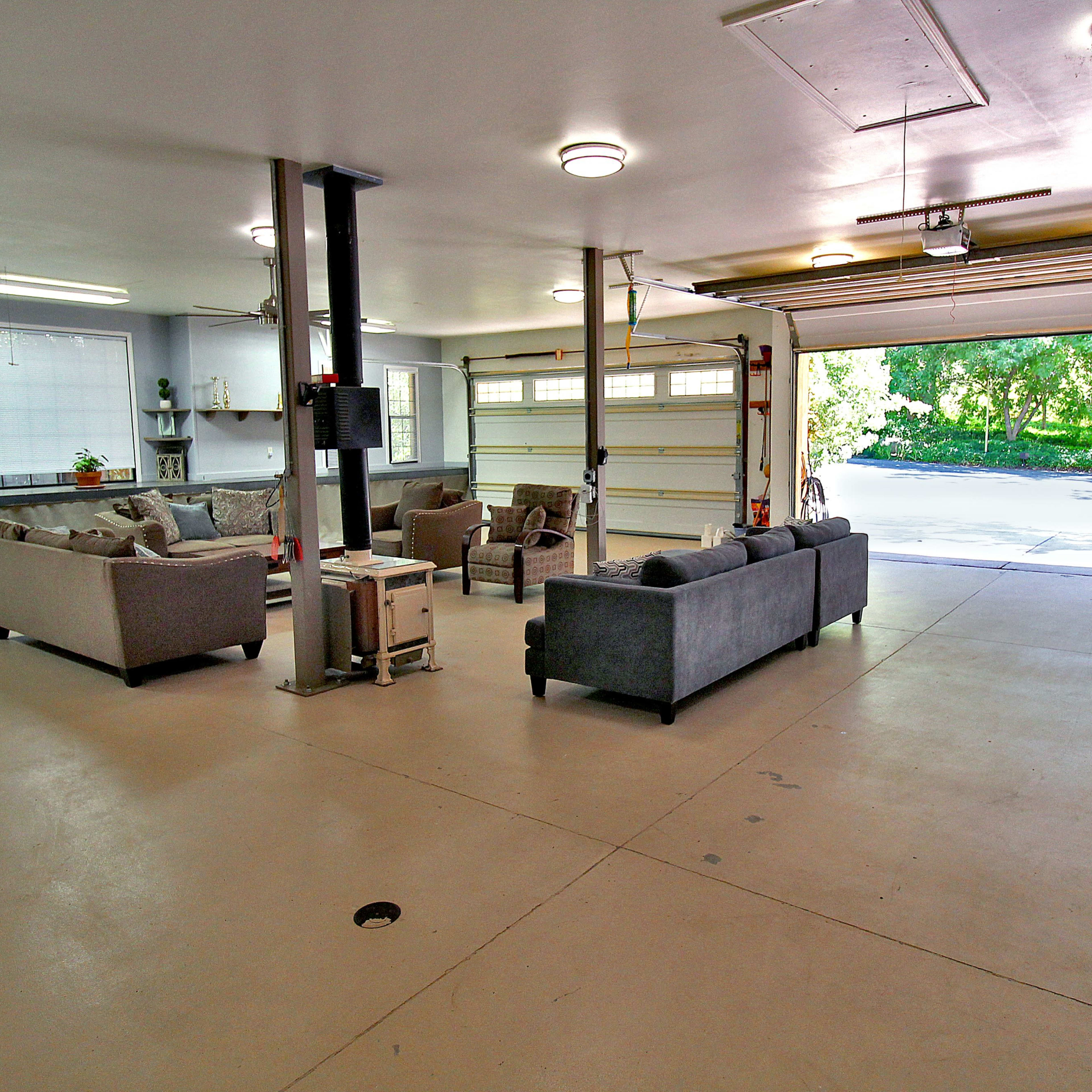 A spacious garage with two sofas arranged around a small table, and a large open garage door revealing a view of the outdoors.