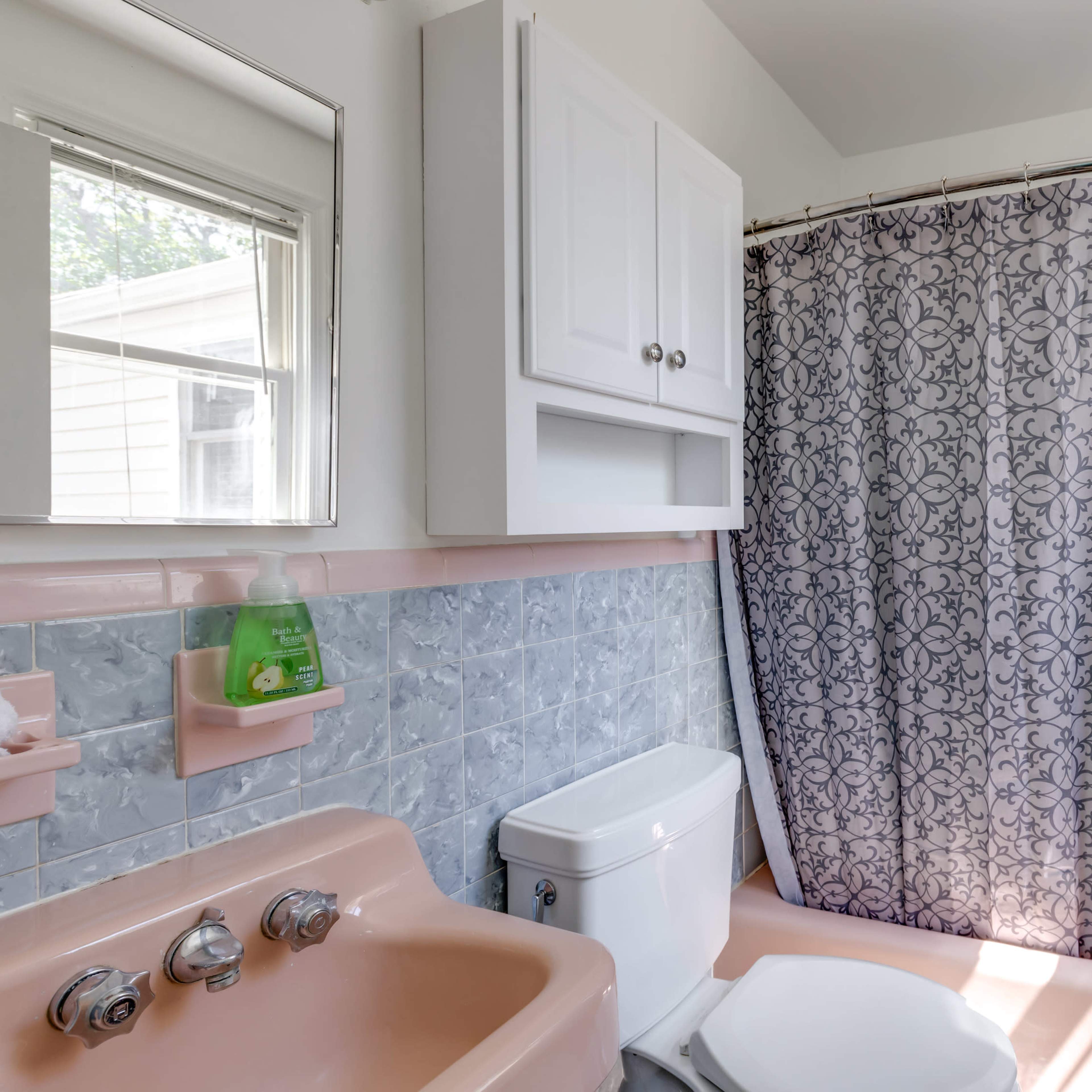 A bathroom with a pink bathtub and toilet, a shower curtain with a patterned design, and a small cabinet above the sink area.