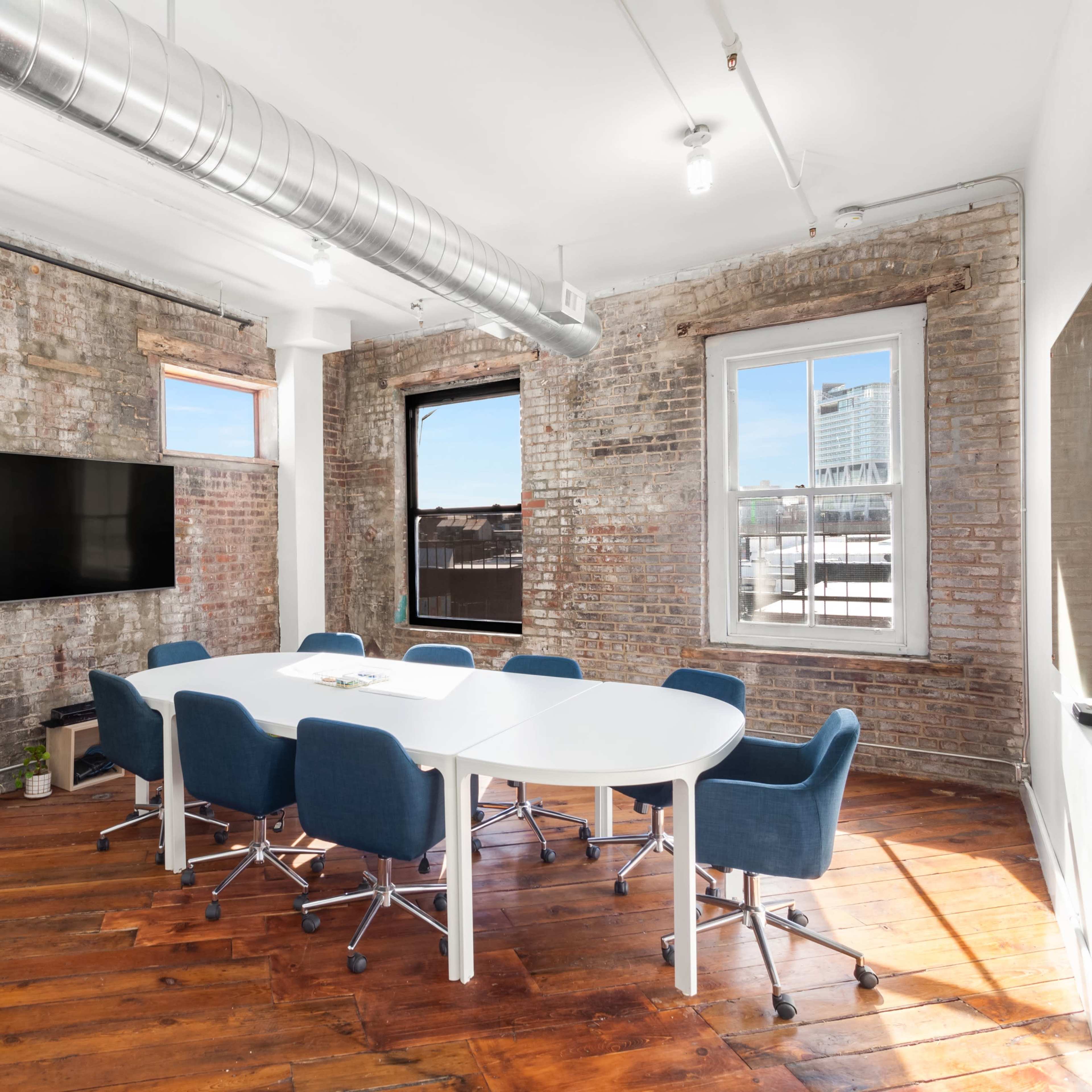 A modern conference room with a white oval table surrounded by blue chairs, exposed brick walls, and windows allowing natural light.