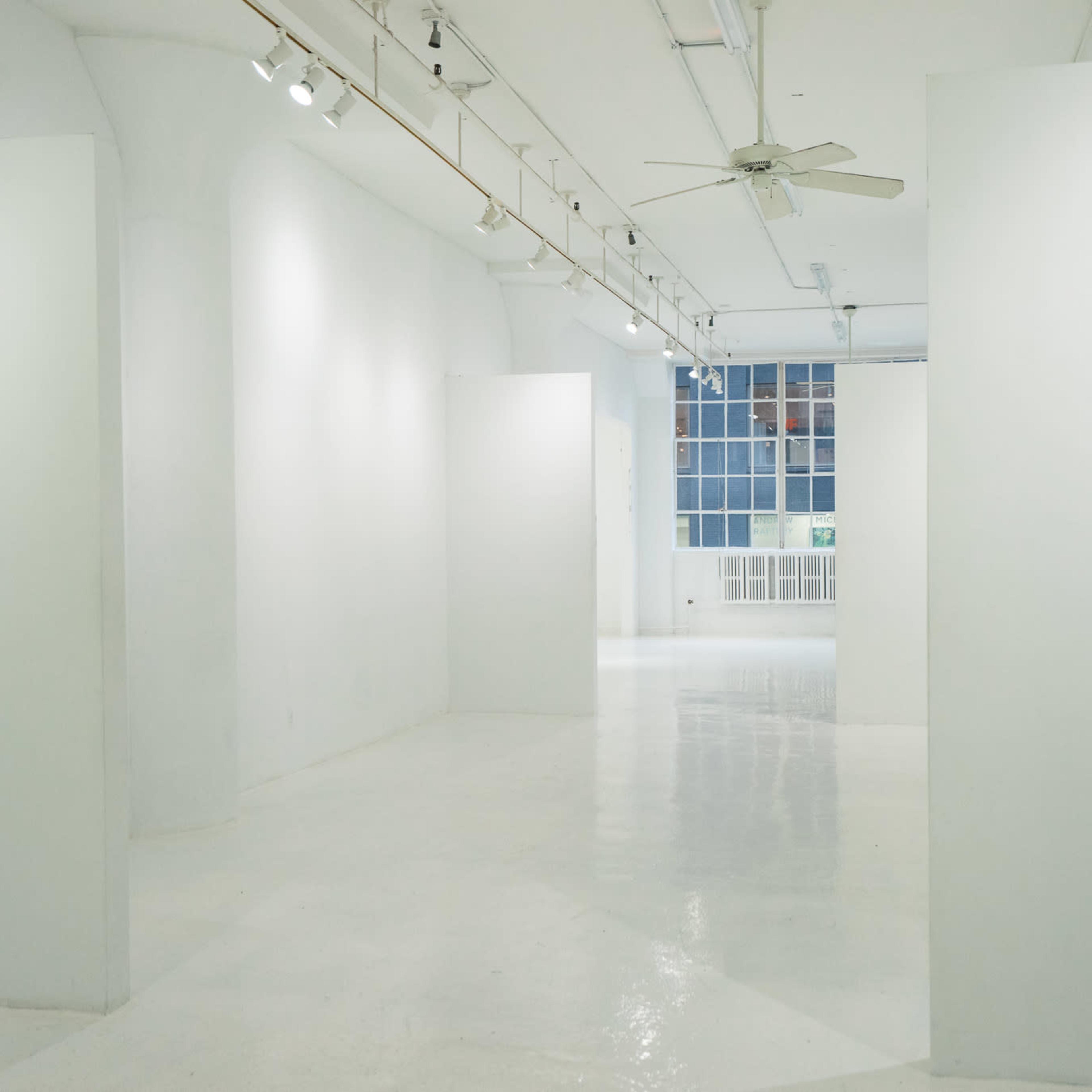 A spacious, white-walled art gallery featuring empty exhibition spaces and natural light coming through large windows.