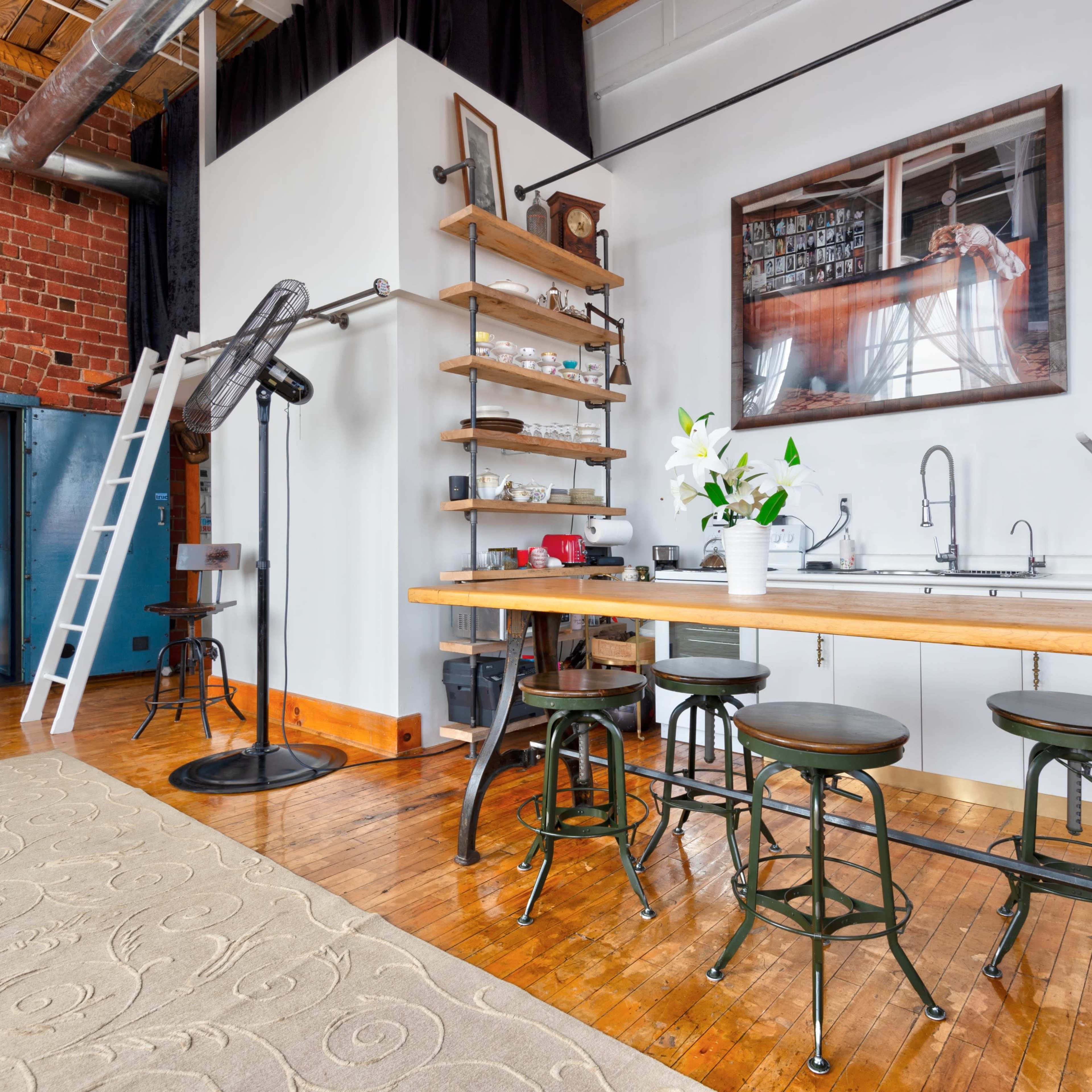 A modern kitchen space with a wooden table, stools, and a bright red refrigerator in an industrial-styled room featuring brick walls and a ladder leading to a loft area.