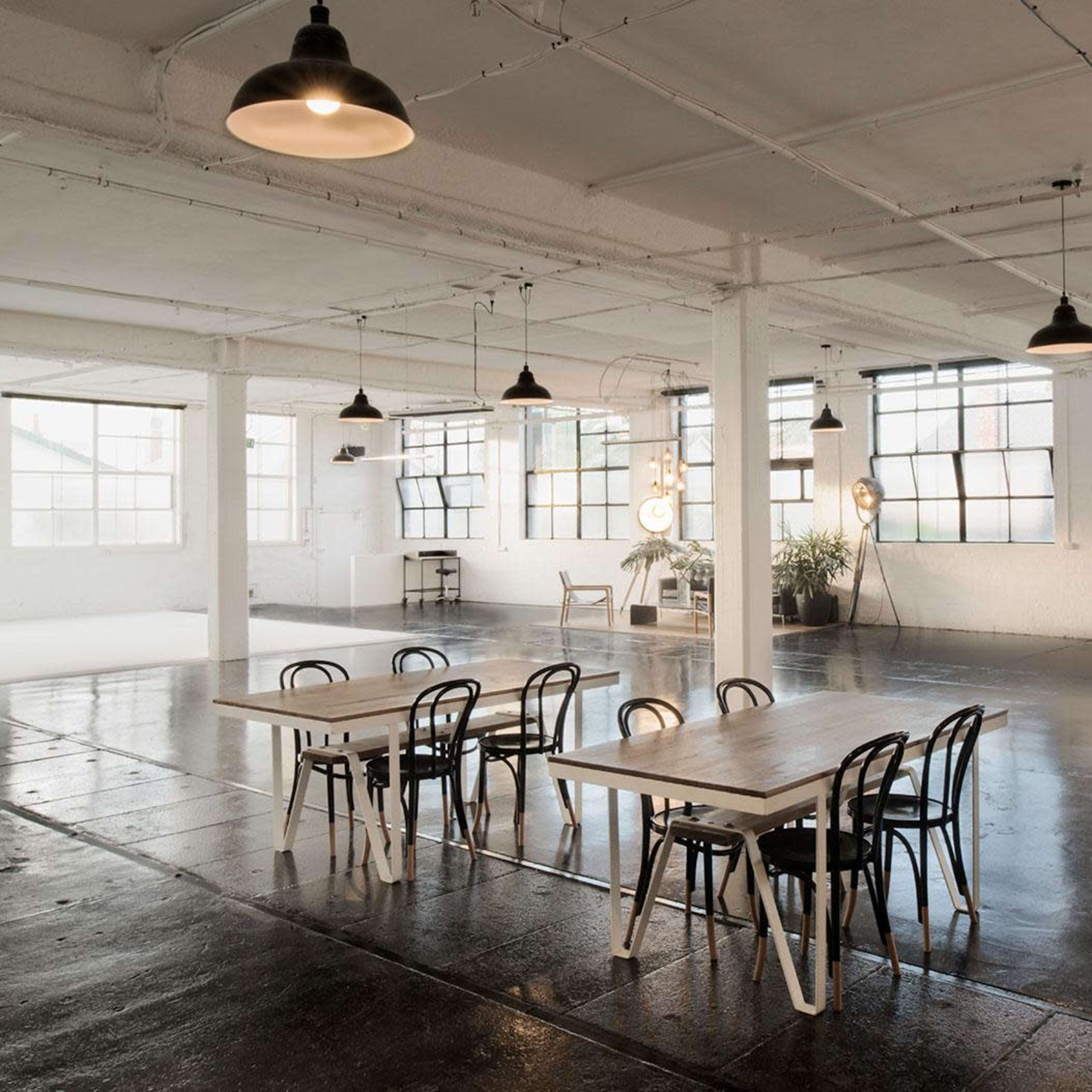 An industrial-style room with polished concrete floors, large windows, and several wooden tables and chairs arranged for a creative workspace.