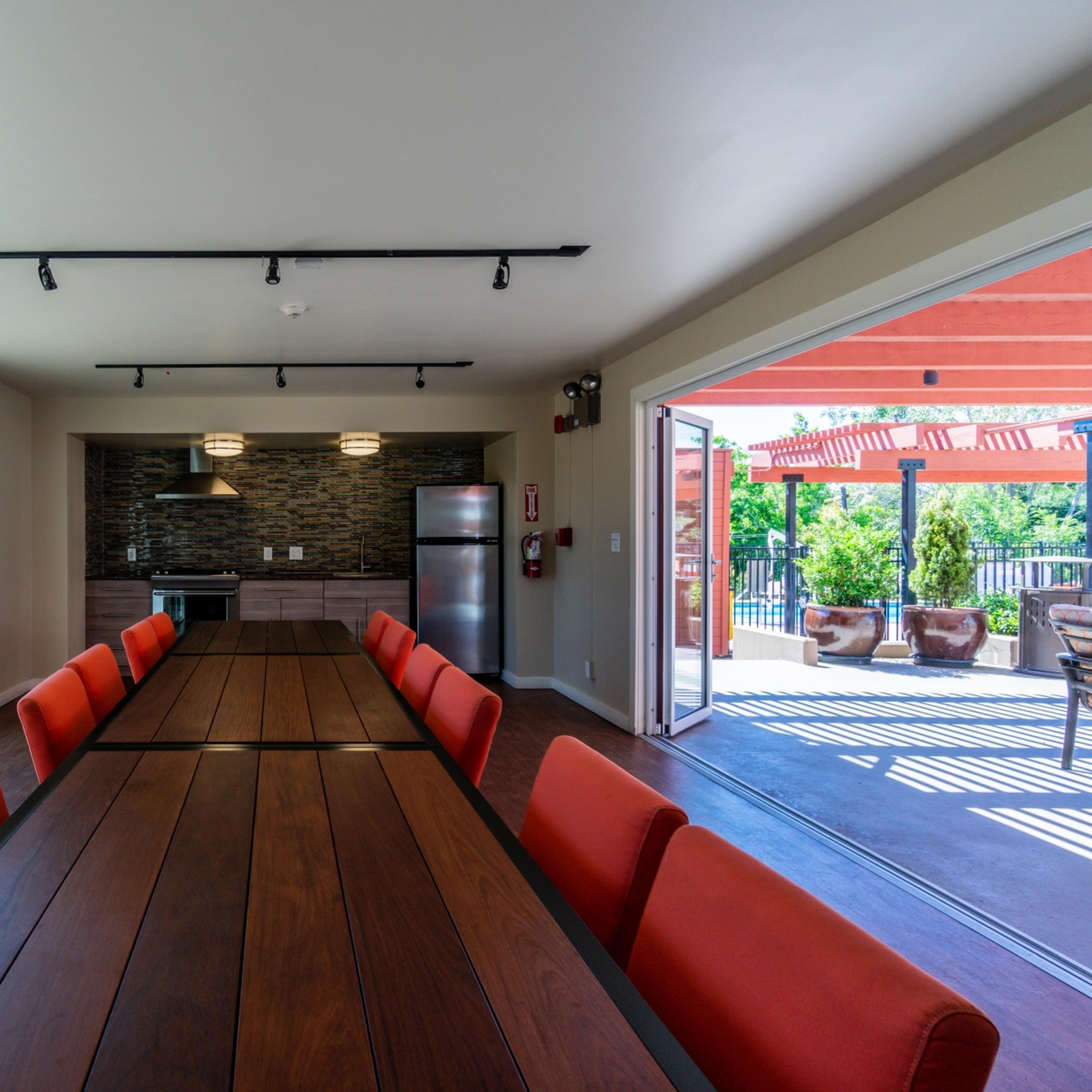 A conference room with a long wooden table surrounded by red chairs, large windows allowing natural light, and a kitchen area in the background.