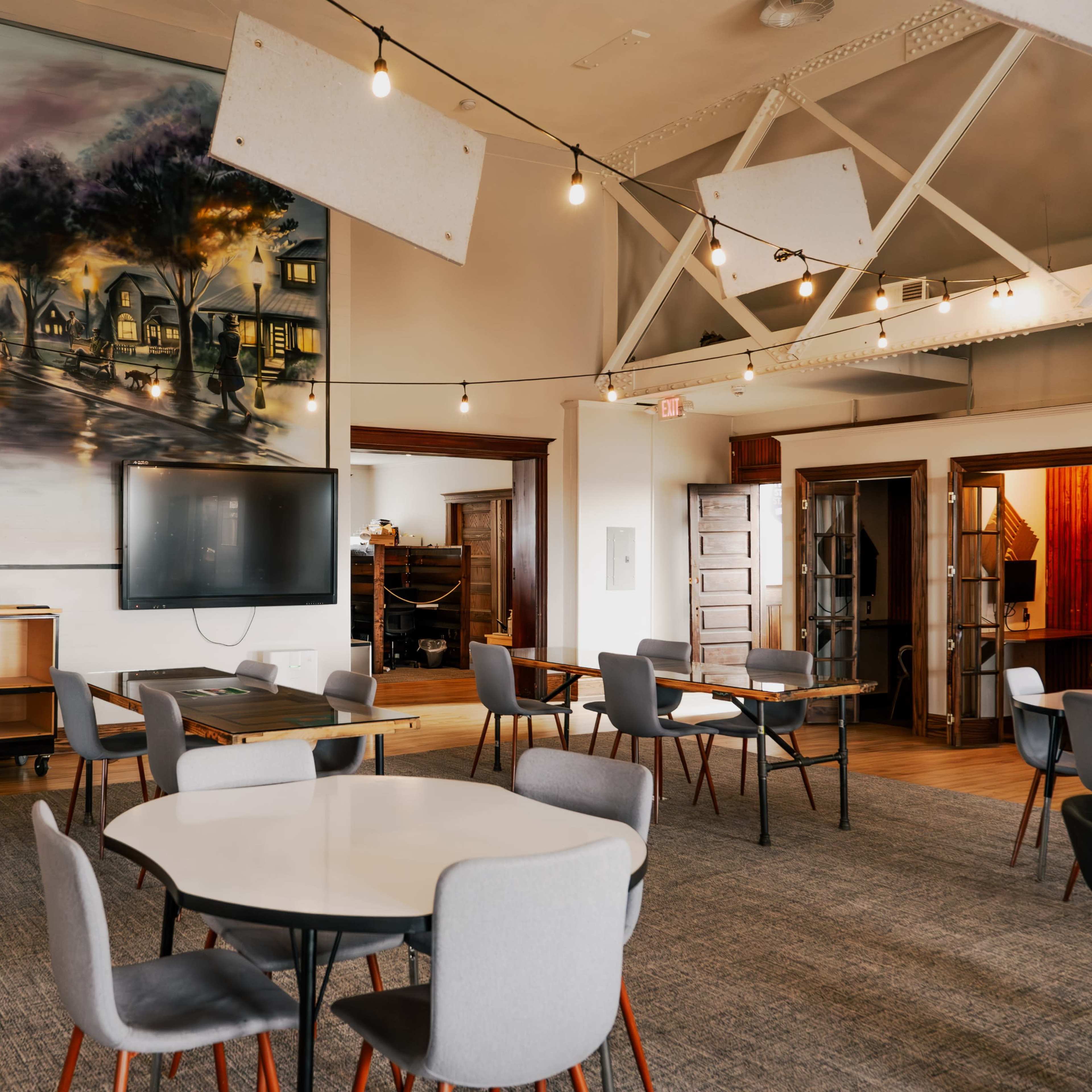 The interior of a room features several round tables with chairs, a large mural on the wall, and a television mounted on a cabinet.