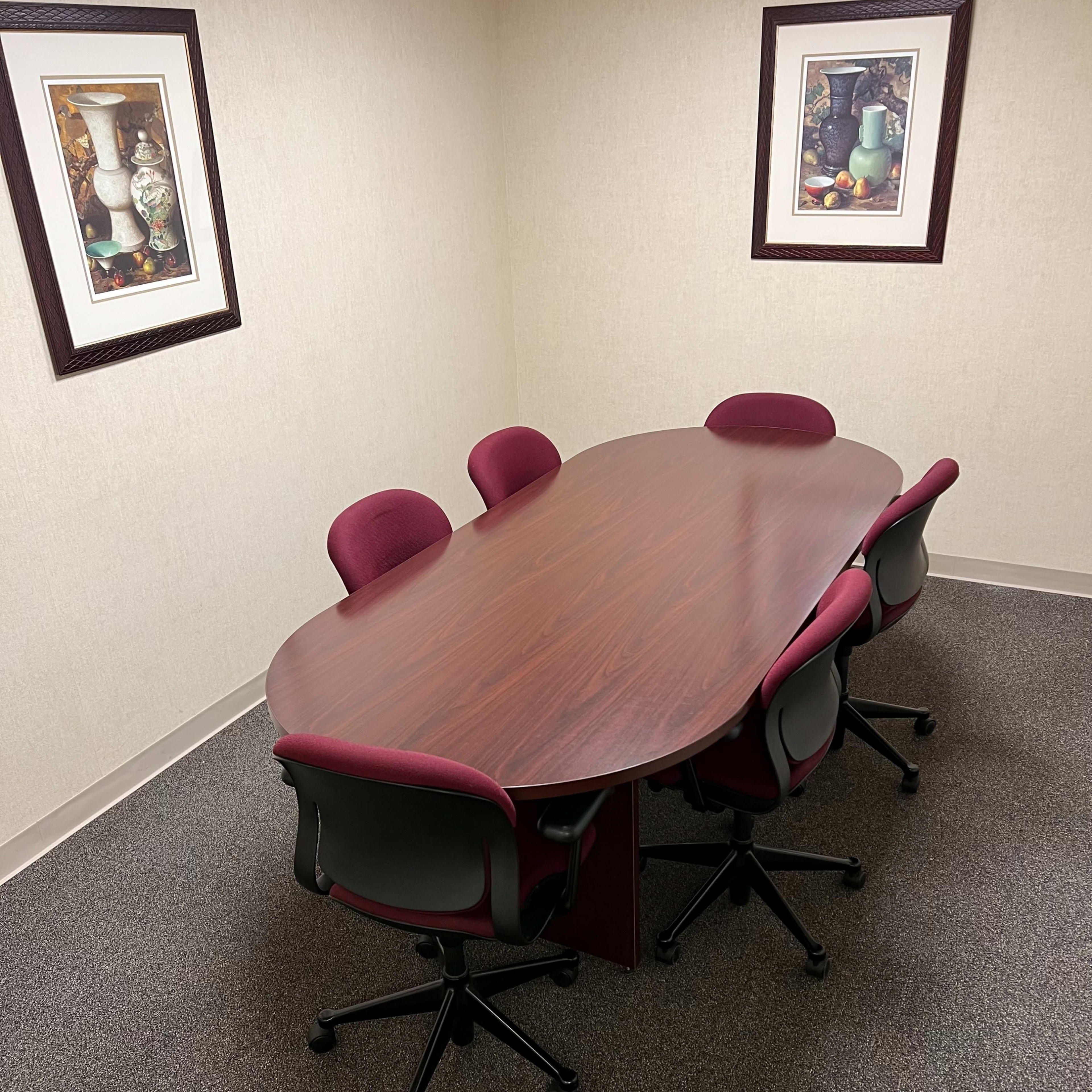 A wooden oval conference table with five burgundy chairs is placed in a small, carpeted meeting room adorned with two framed artwork pieces on the walls.