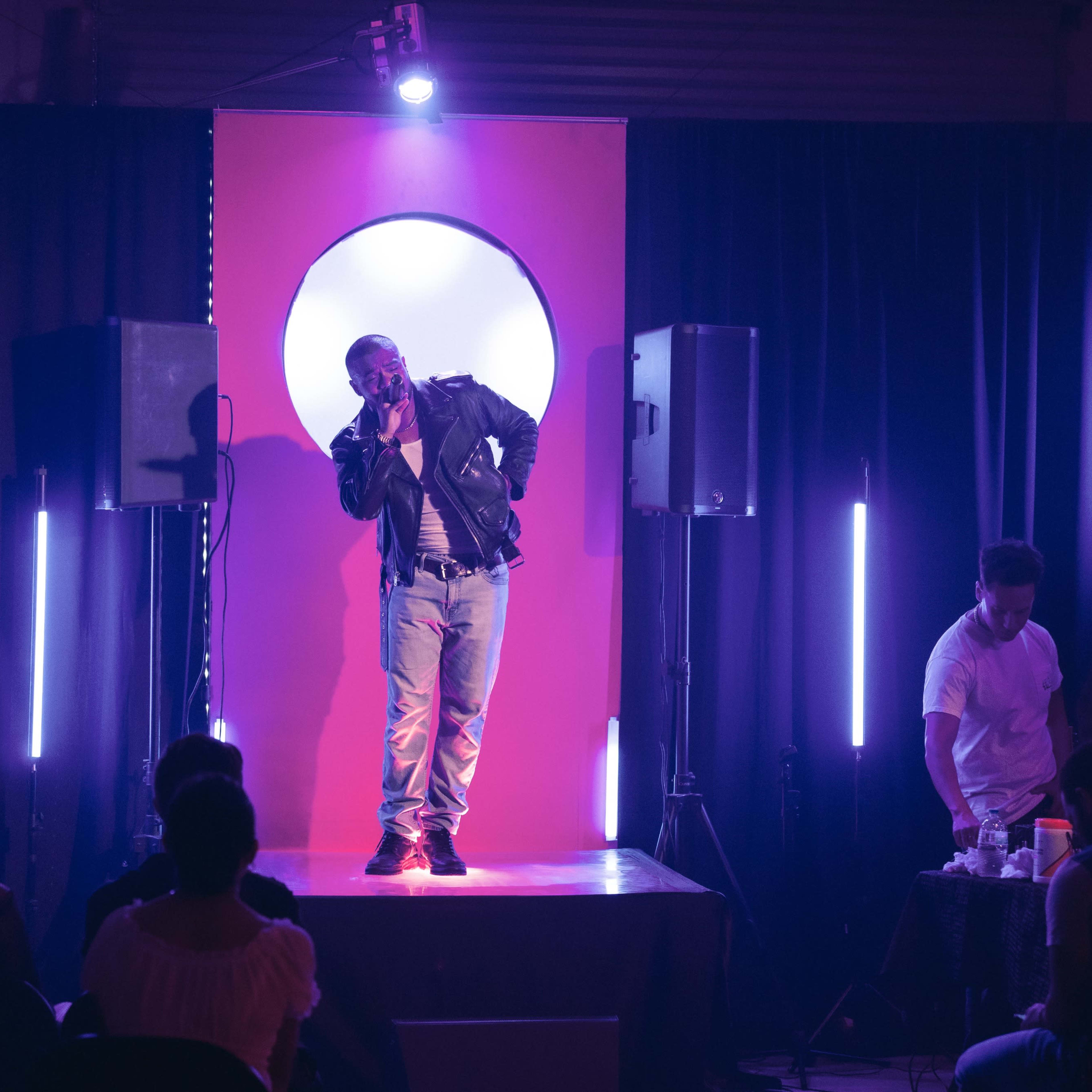 A performer stands on stage in front of a circular backdrop, delivering a routine as the audience looks on.