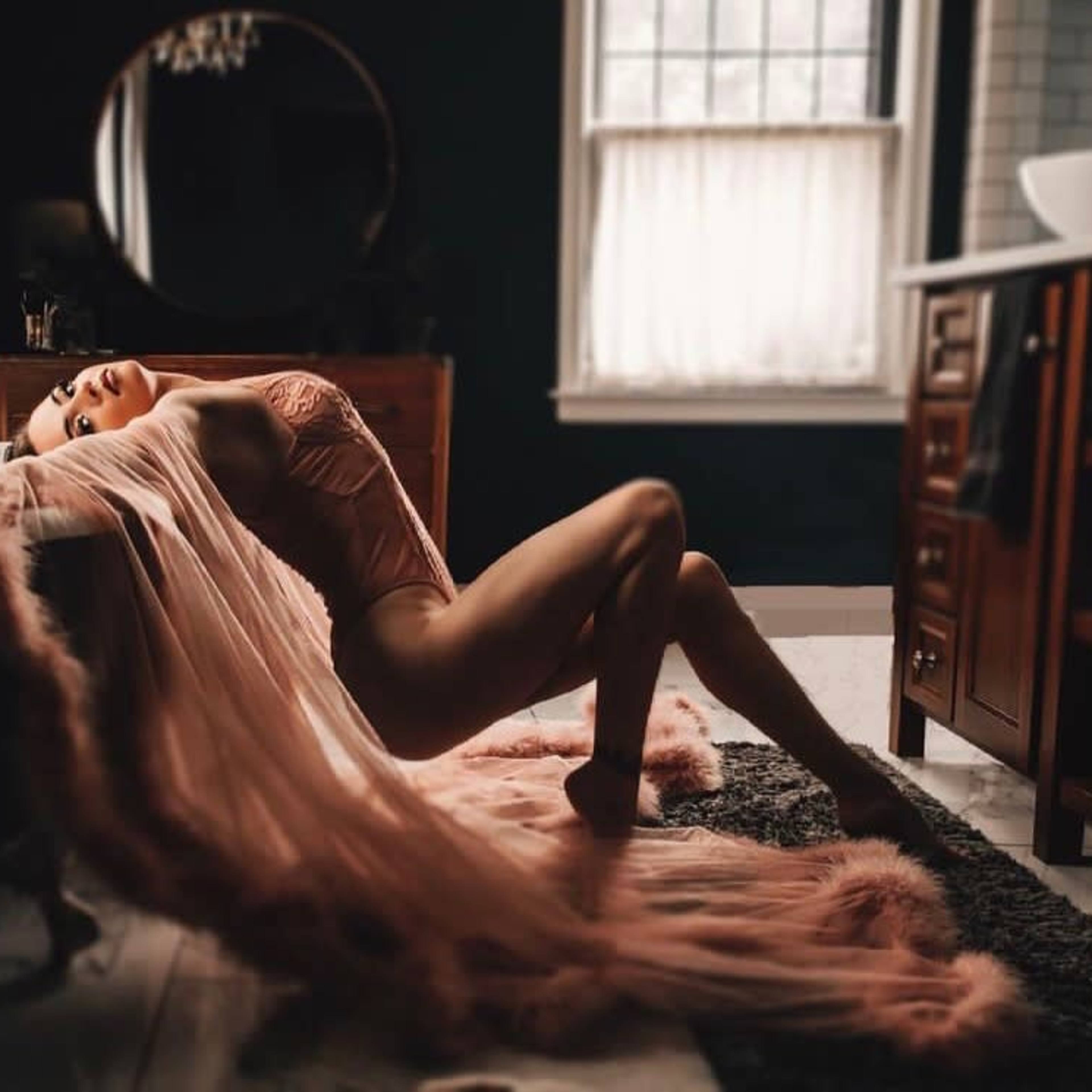 A person in a pink garment poses gracefully near a bathtub, draped with a long, flowing fabric in a dimly lit bathroom.