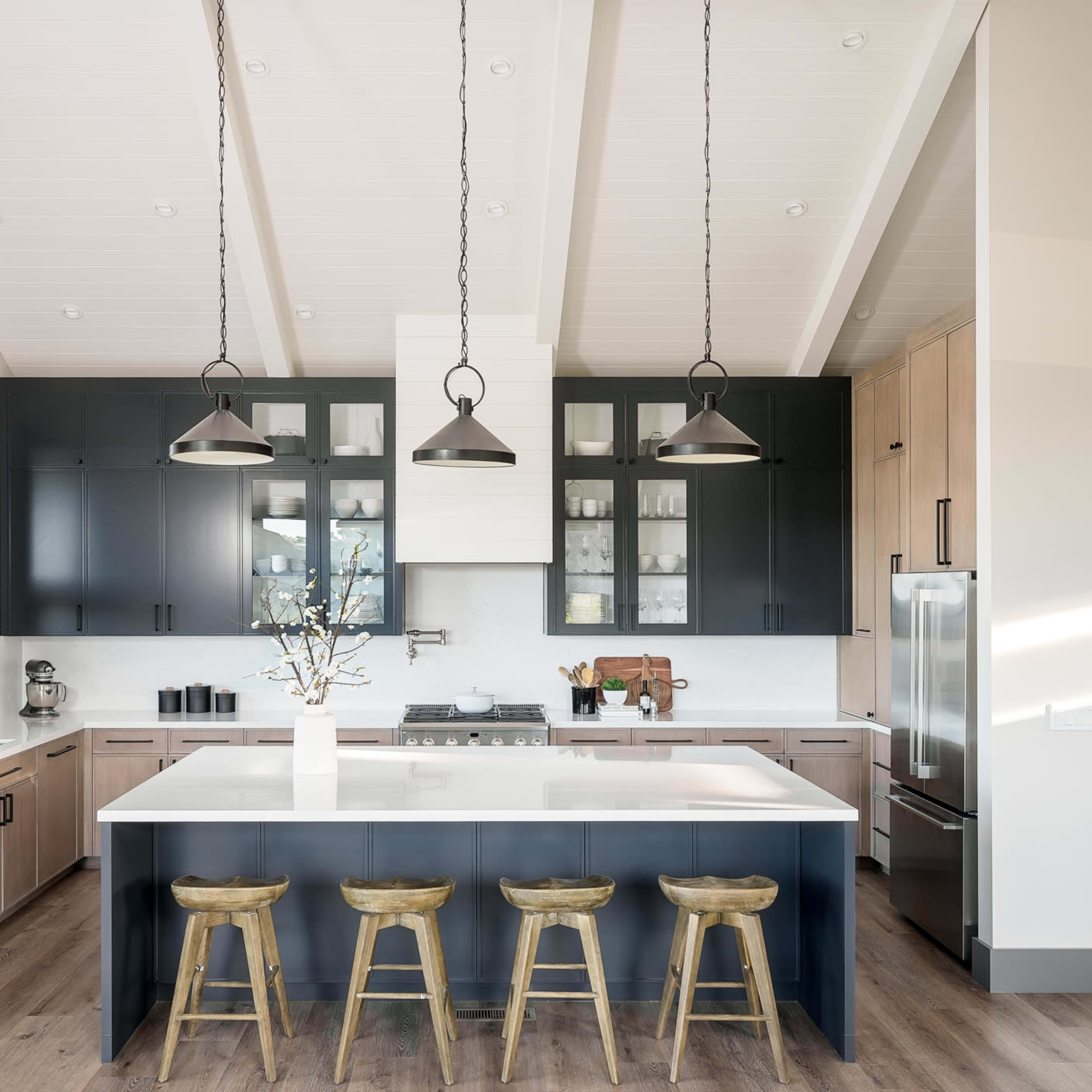 A modern kitchen featuring a large island with barstools, dark cabinetry, and a light countertop, complemented by pendant lighting and wooden flooring.