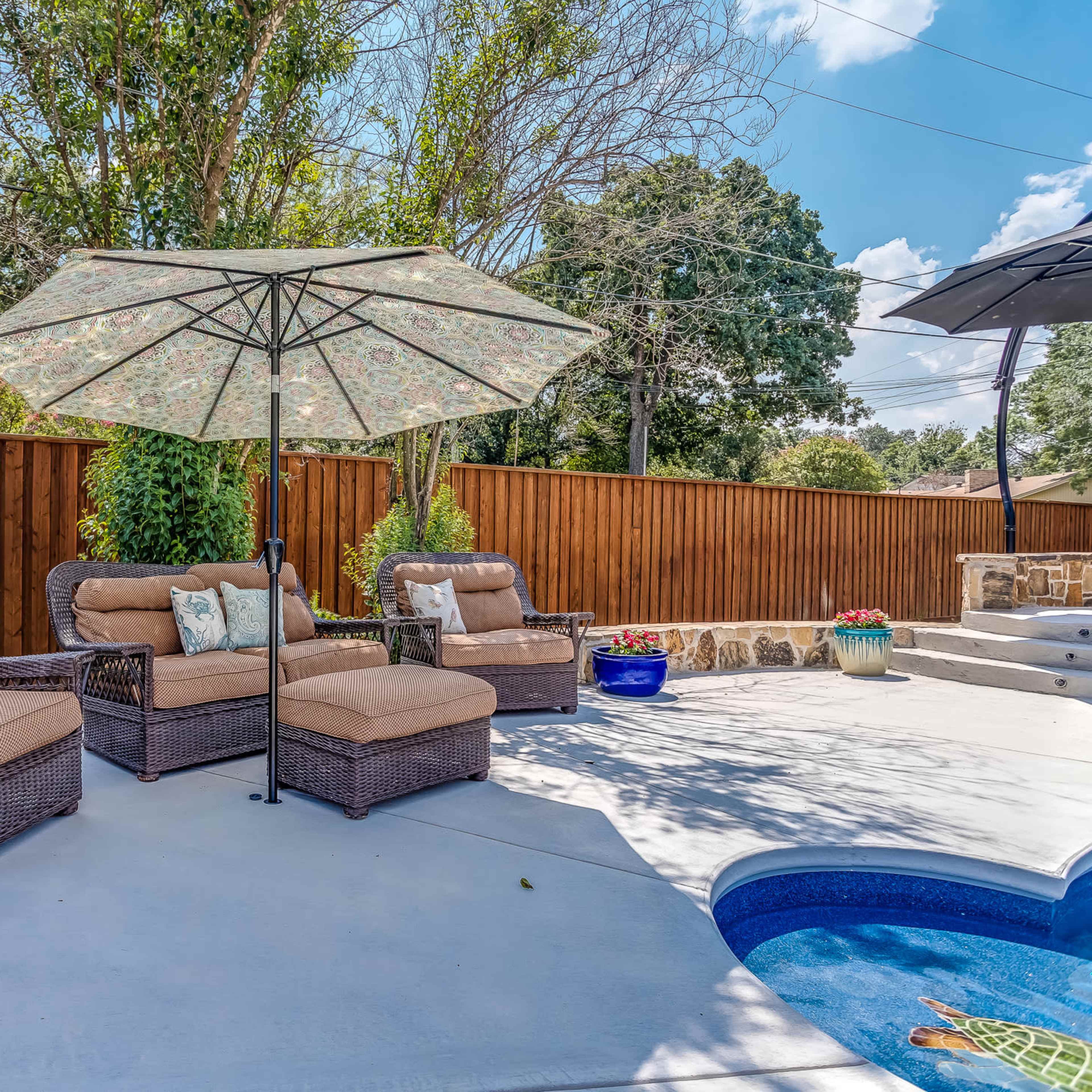 A patio area features wicker chairs with cushions and a large umbrella, surrounded by a wooden fence and trees.