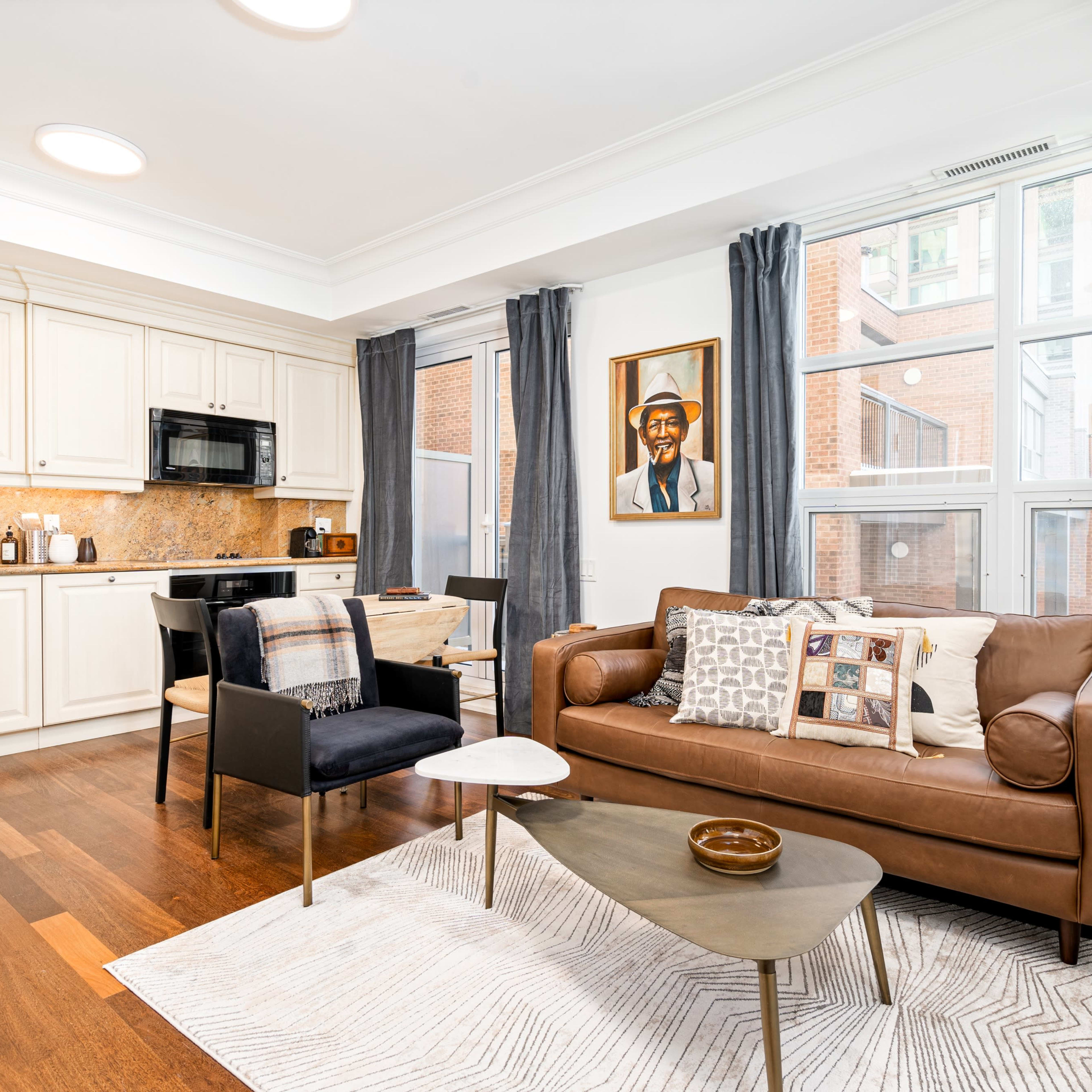 A modern apartment living area featuring a kitchen with light cabinetry, a sitting area with a brown leather couch and a small coffee table, and large windows providing natural light.
