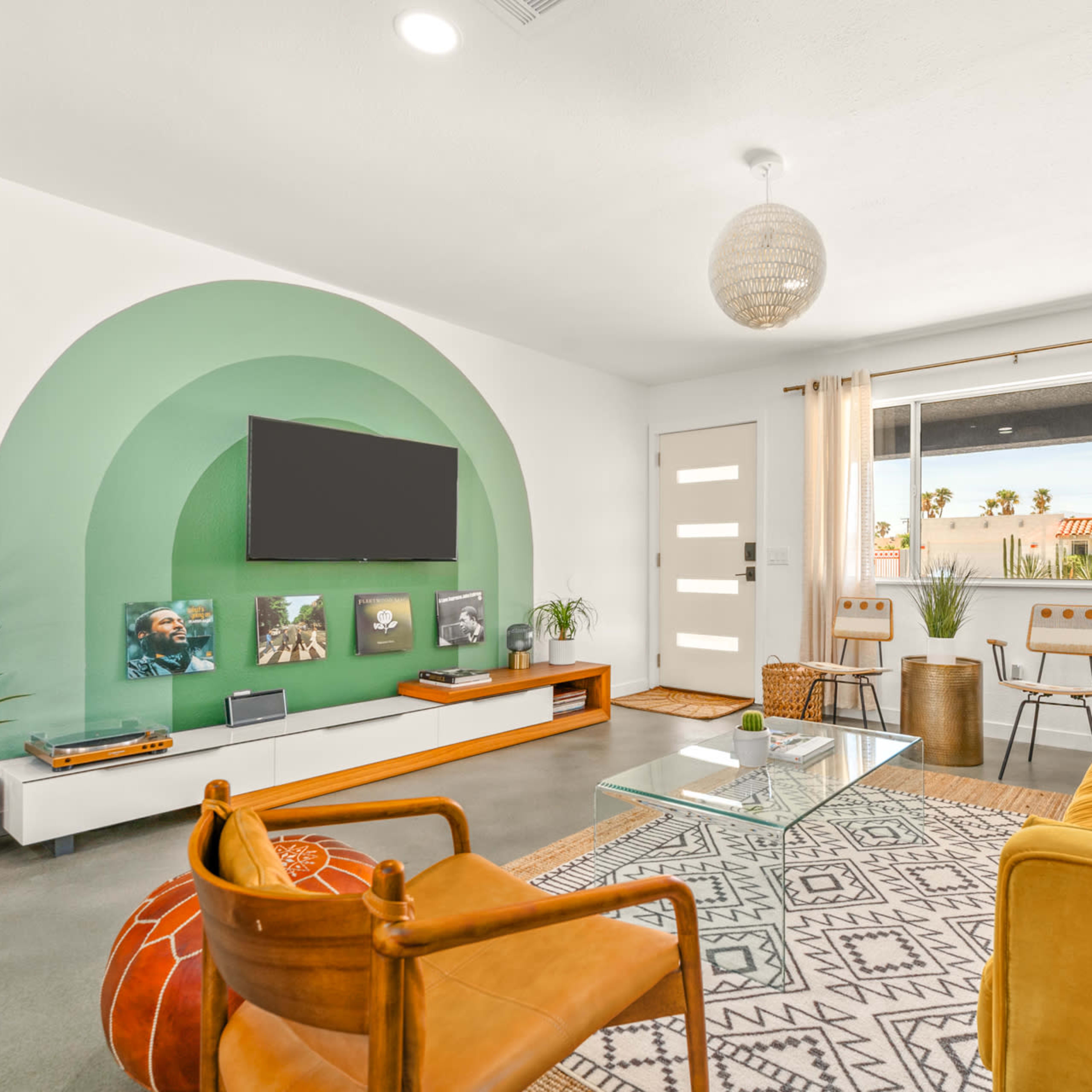 A bright living room featuring a green arch wall design, a large television, a glass coffee table, and modern furniture including a mustard-colored sofa and wooden chairs.