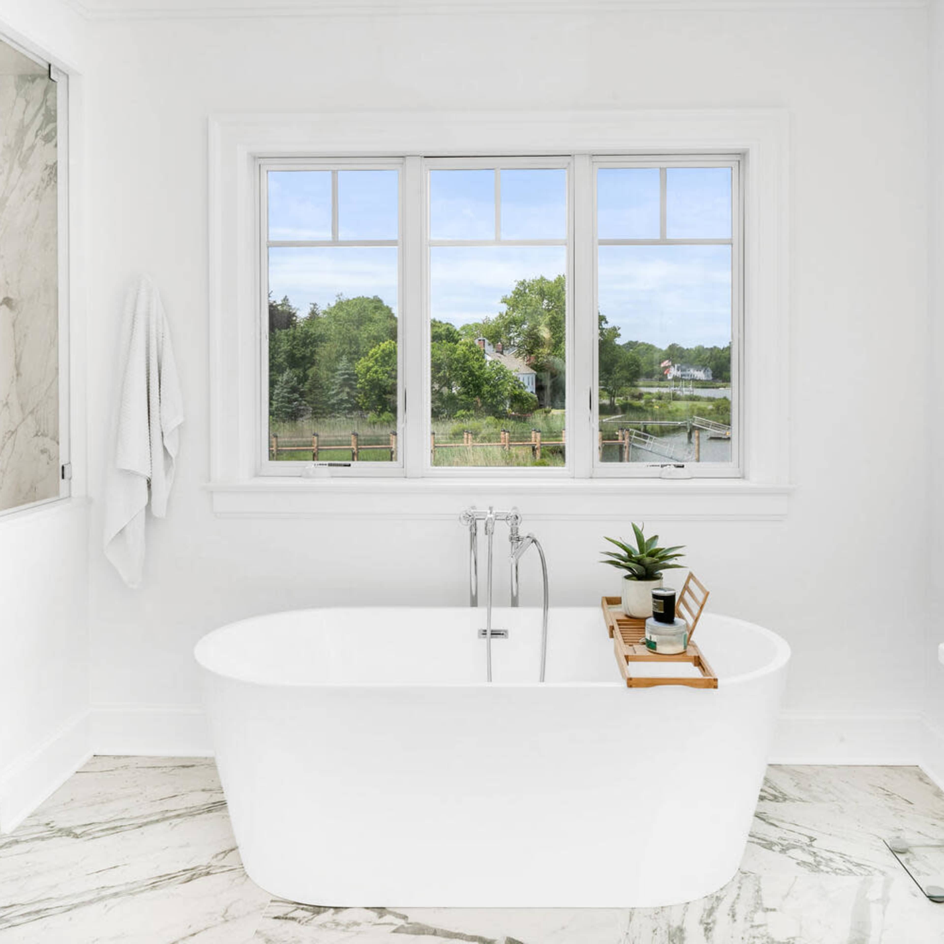 A freestanding white bathtub is positioned in a bright bathroom with large windows overlooking a green landscape.