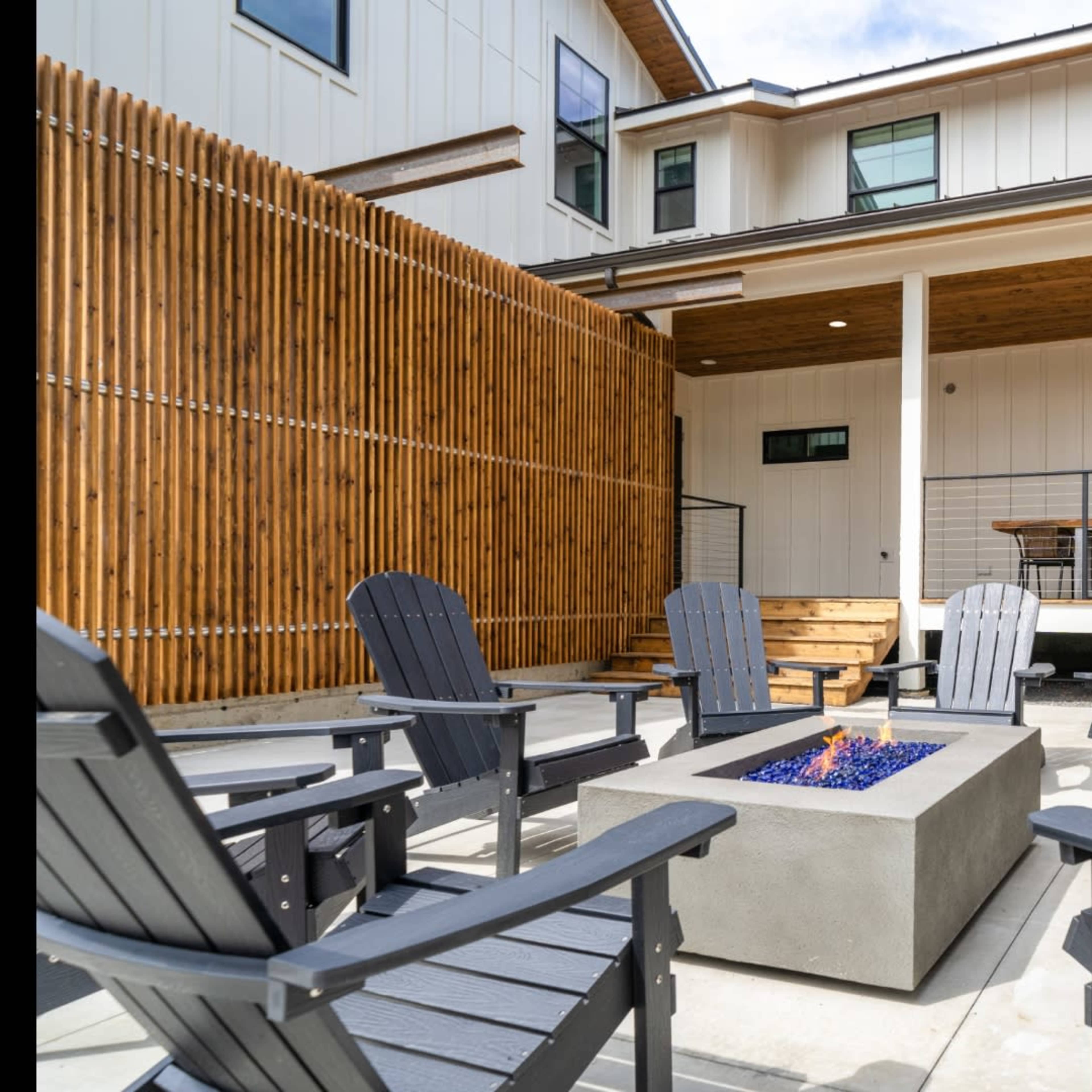A modern outdoor seating area with wooden fencing, a gas fire pit, and several gray Adirondack chairs arranged around it.