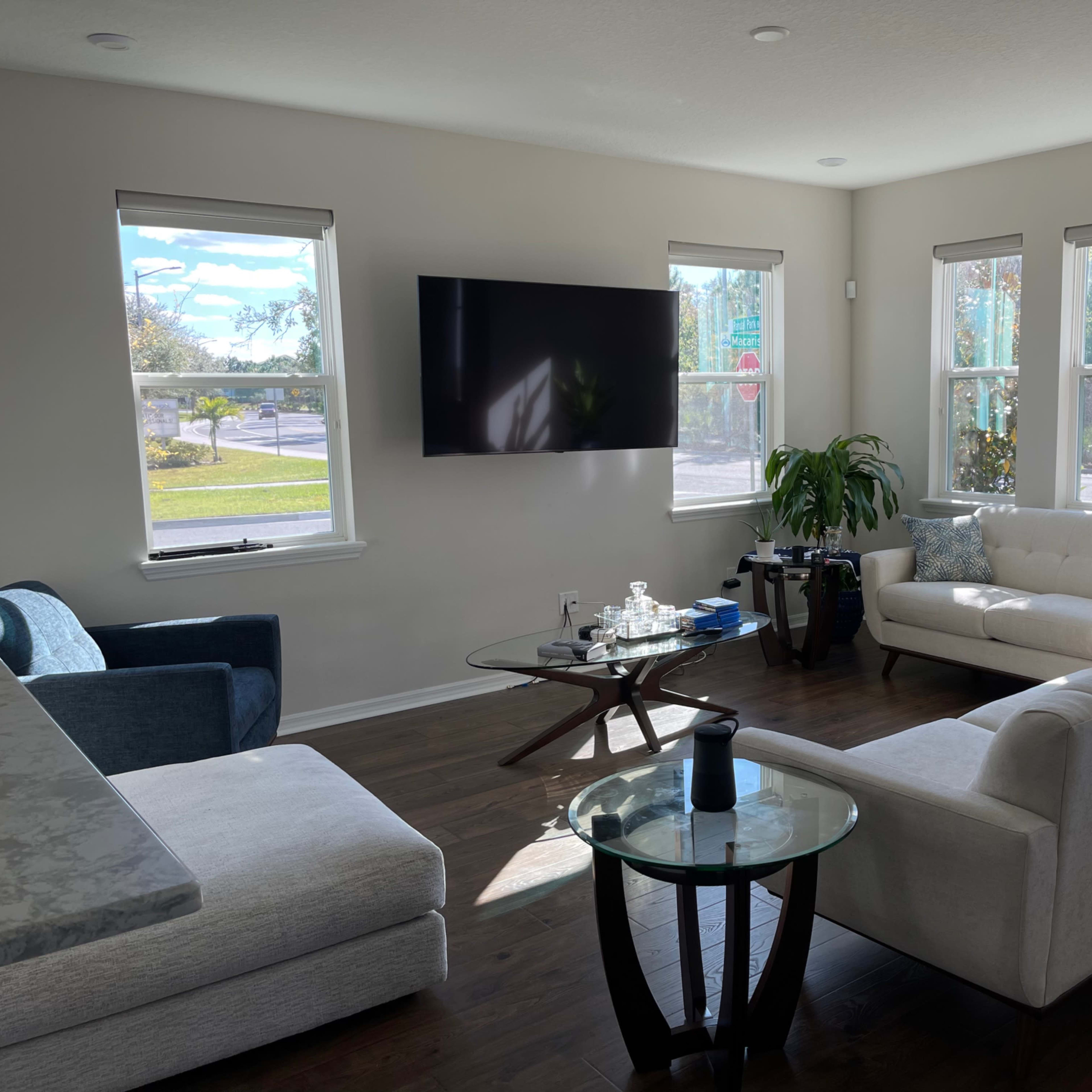 A living room features a white sofa, a blue chair, a glass coffee table, a TV mounted on the wall, and several windows allowing natural light to enter.