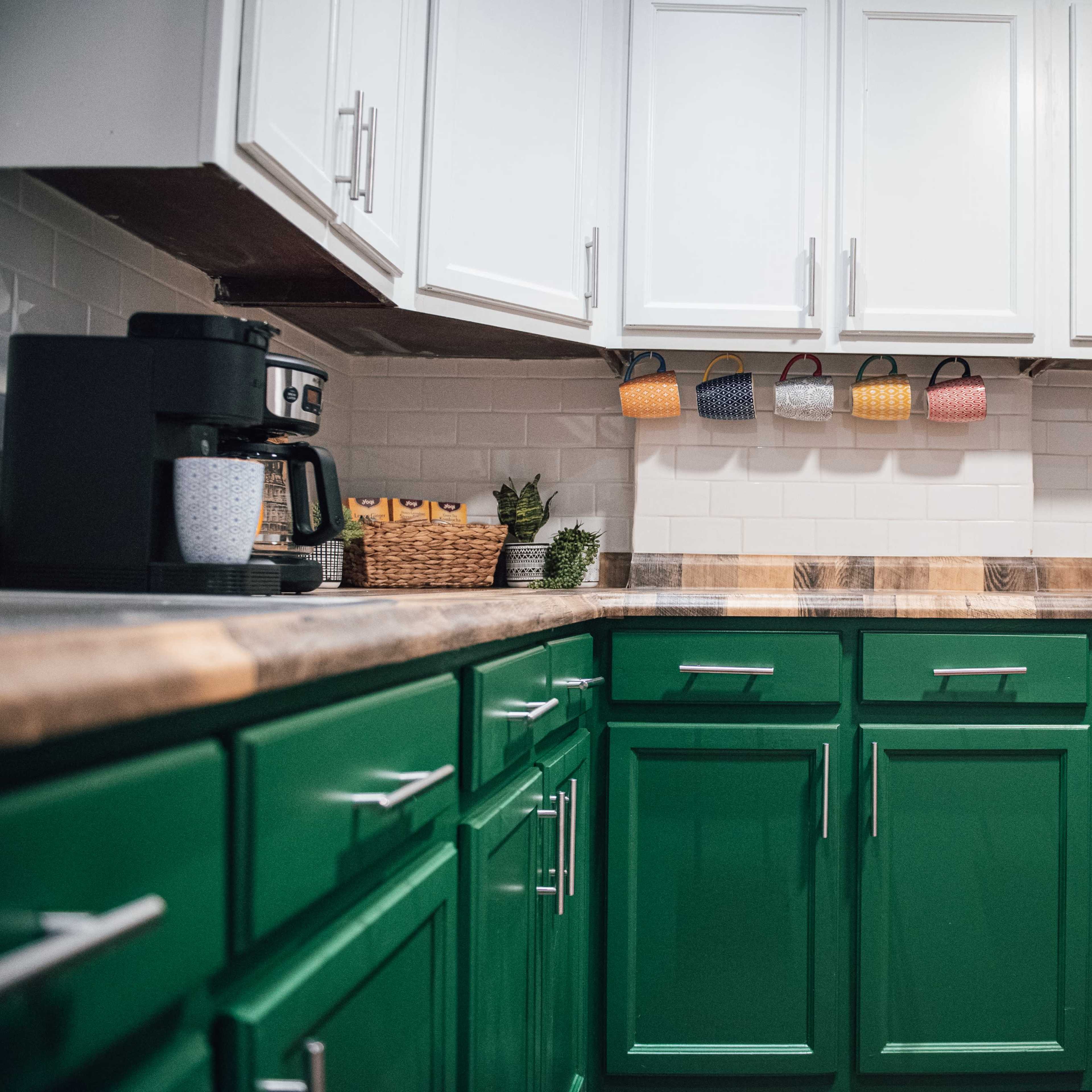 A modern kitchen with green lower cabinets, white upper cabinets, and a countertop made of wood, featuring a coffee maker and a yellow teapot.
