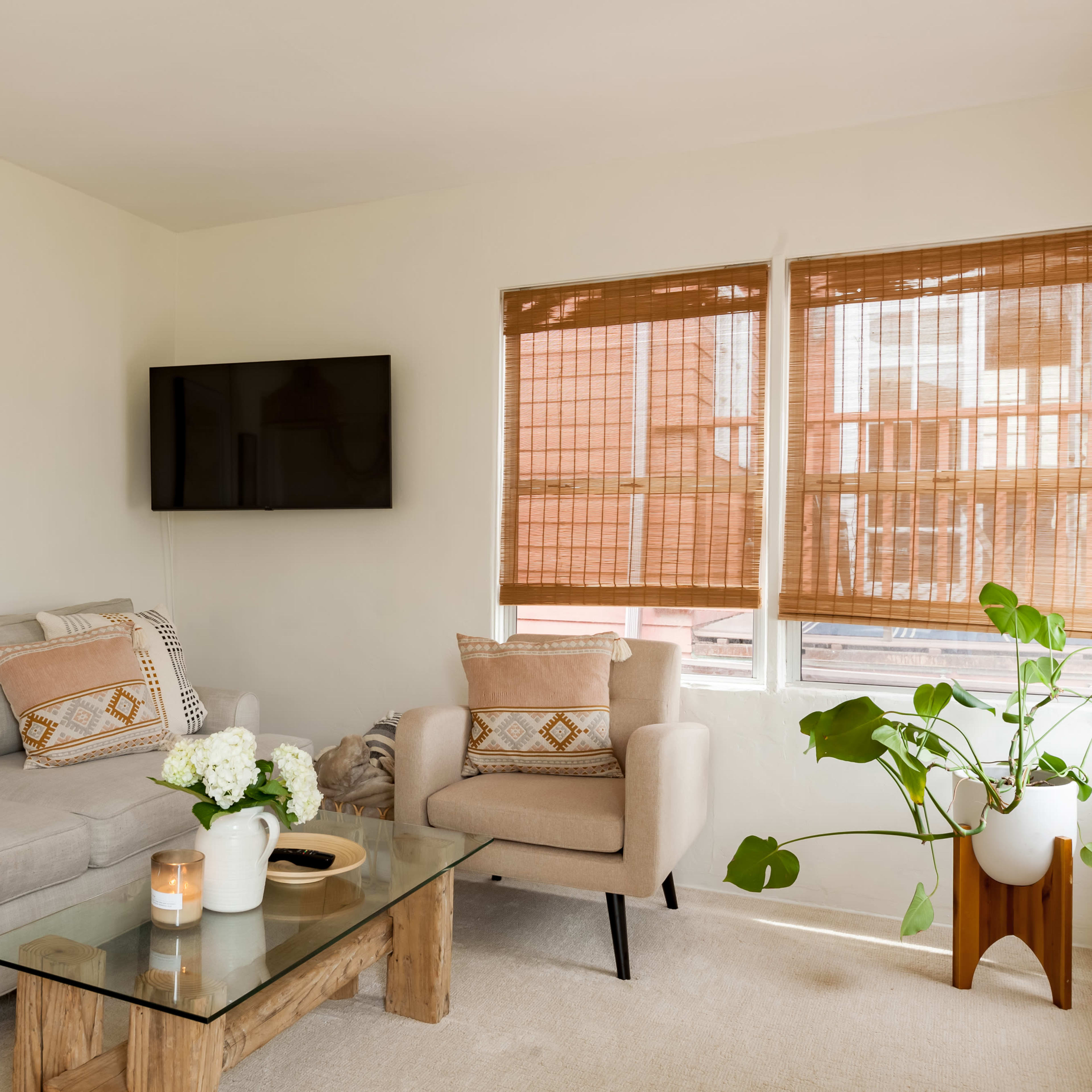 A cozy living room features a light gray sofa, two beige armchairs, a glass coffee table, a television on the wall, and large windows with bamboo shades.