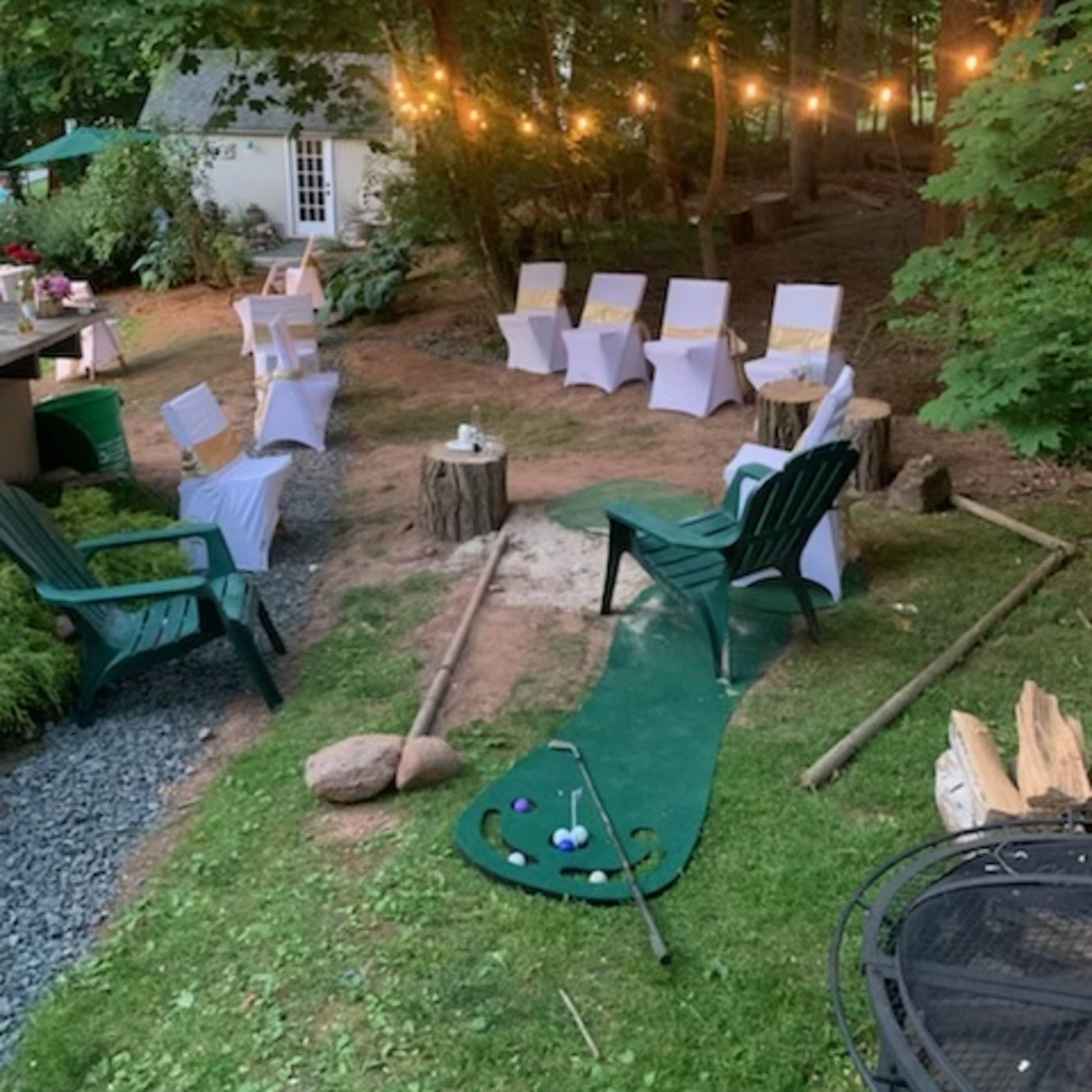 The scene shows a landscaped outdoor area with lawn chairs, string lights, a putting green, and a small table among trees.