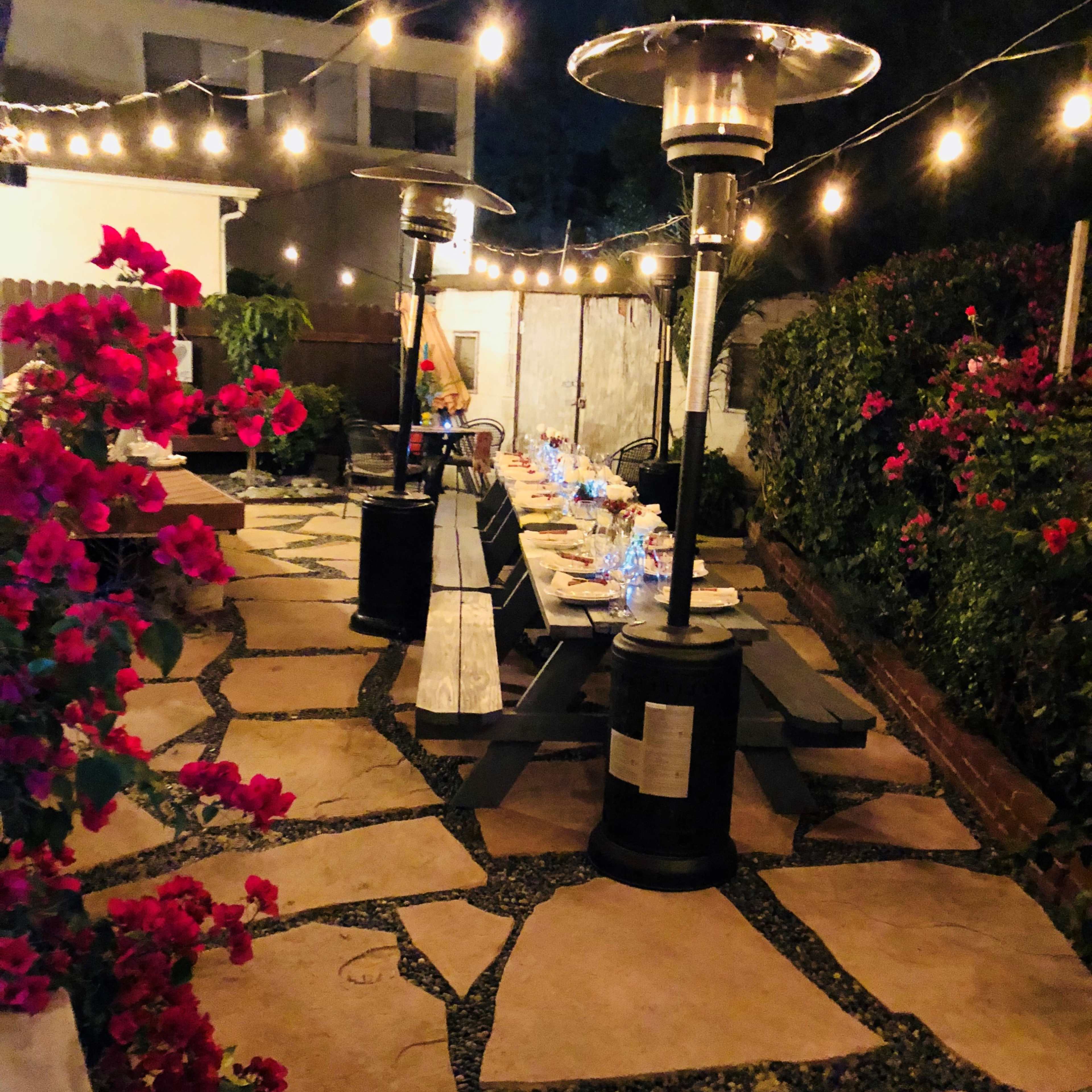 A softly lit outdoor patio area with stone pathways, patio heaters, a long dining table set with glasses, and blooming bougainvillea plants along the edges.