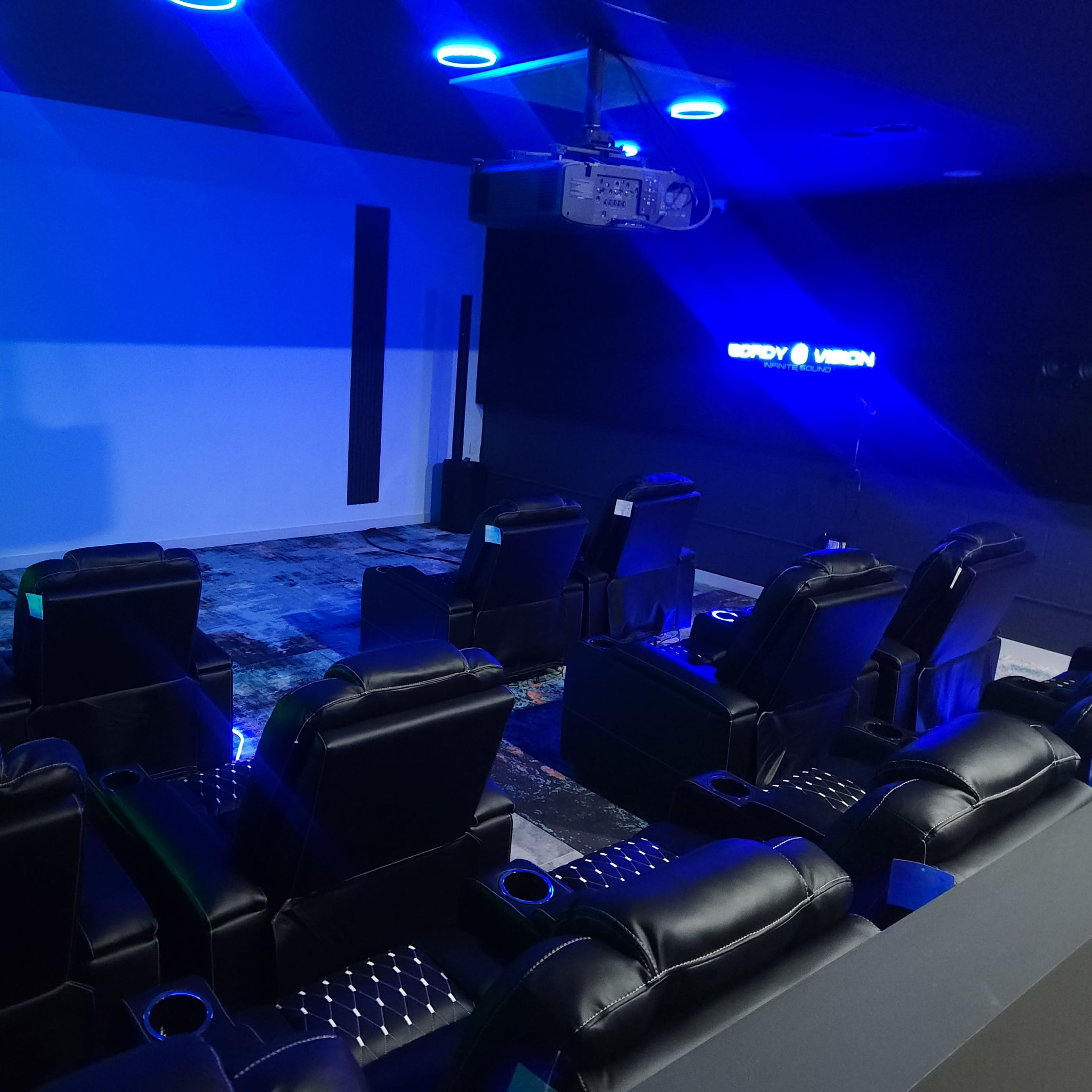 A modern home theater featuring black leather reclining chairs arranged in front of a large blank screen and illuminated by blue LED lights.