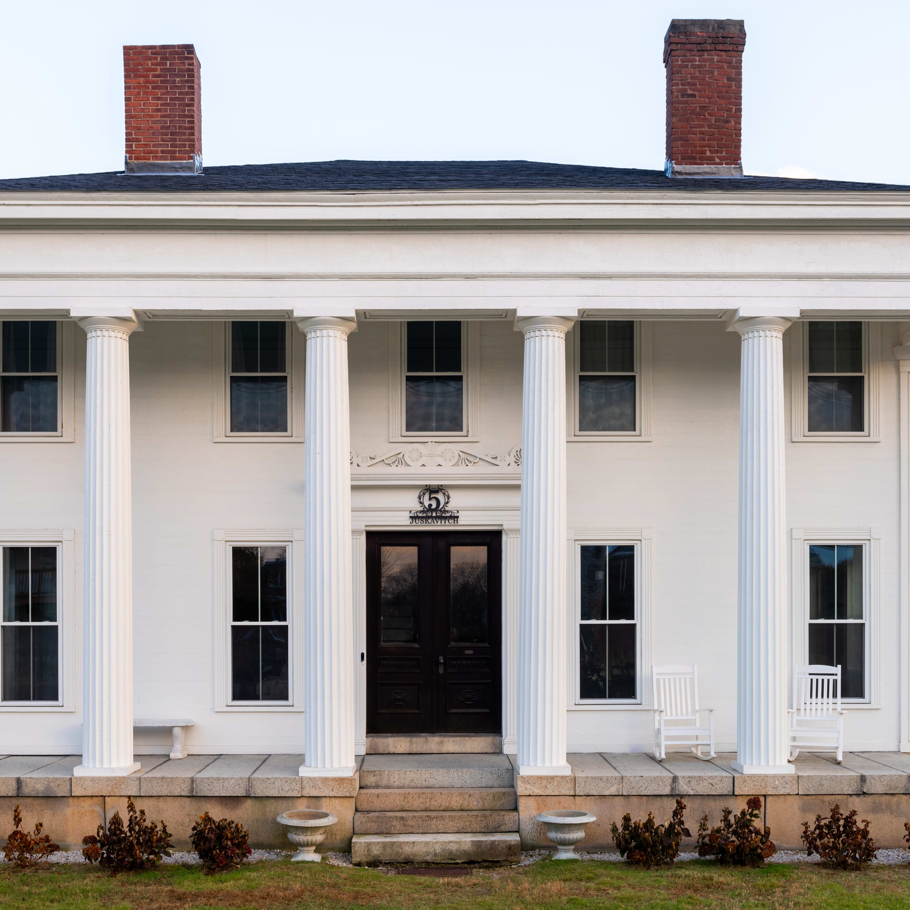 A large, white, classical-style house with columns and a detailed pediment, situated on a grassy area.
