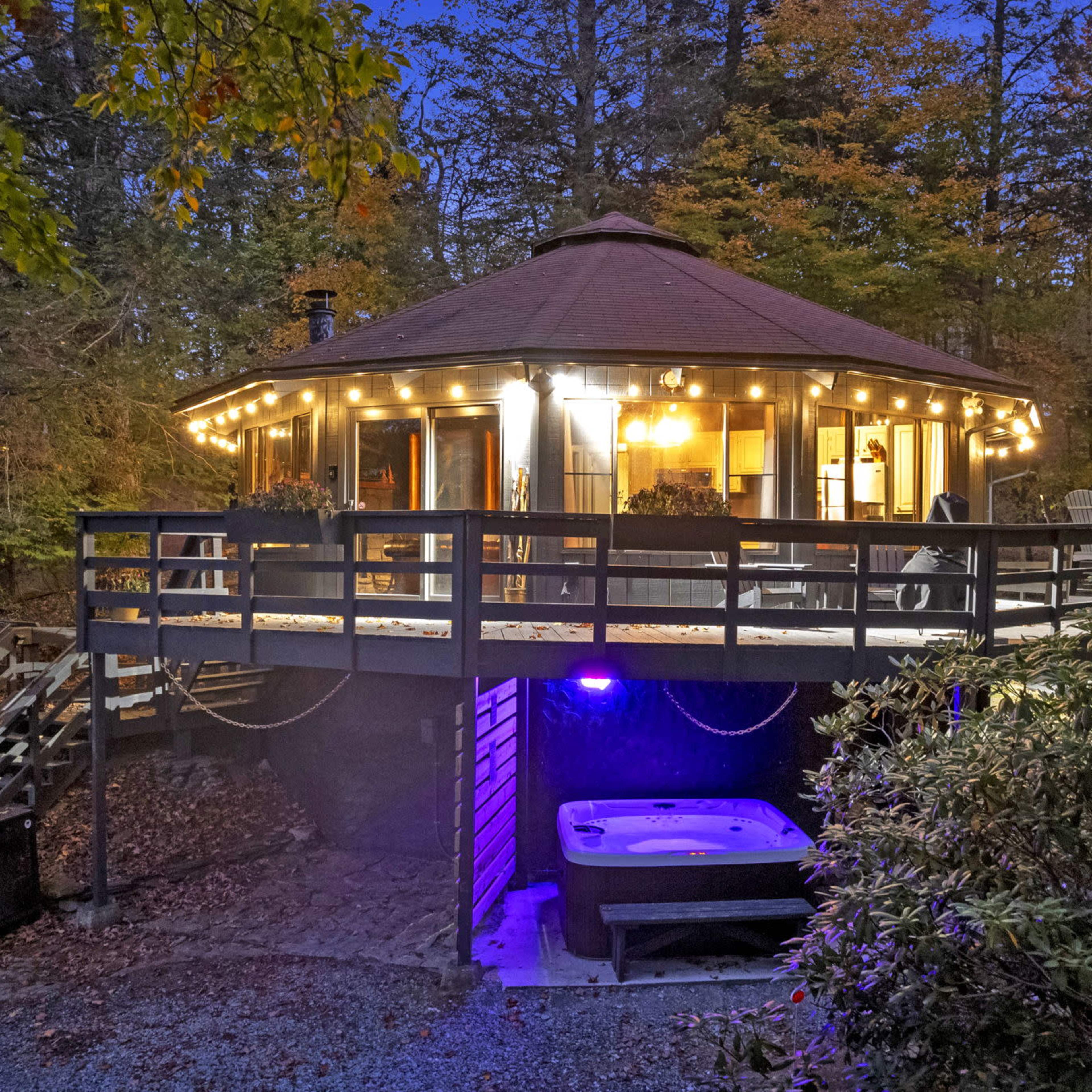 A circular, wooden cabin with a deck is illuminated by string lights and features a hot tub underneath at dusk, surrounded by trees.