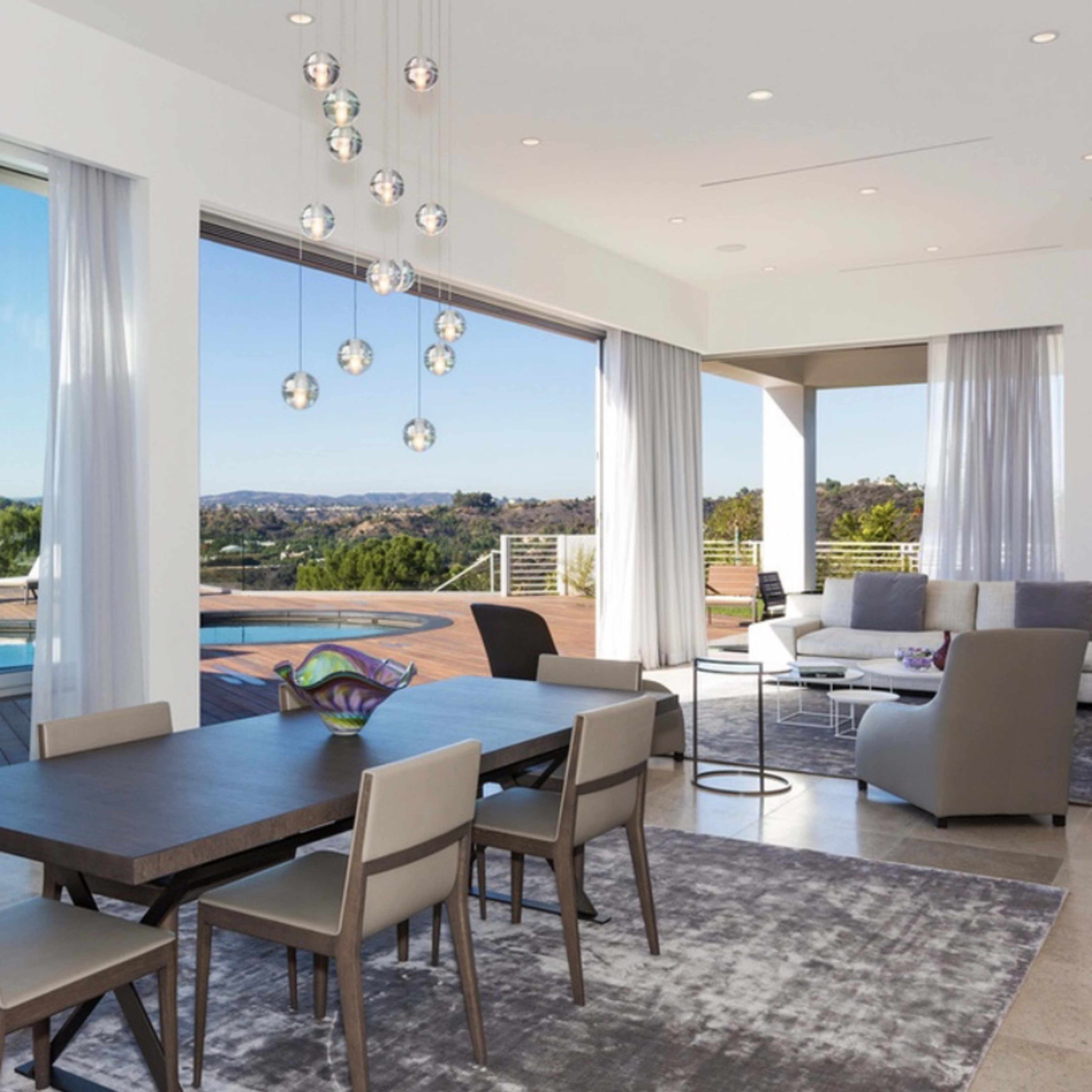 A modern dining area features a large table surrounded by chairs, with expansive windows overlooking a pool and scenic view.