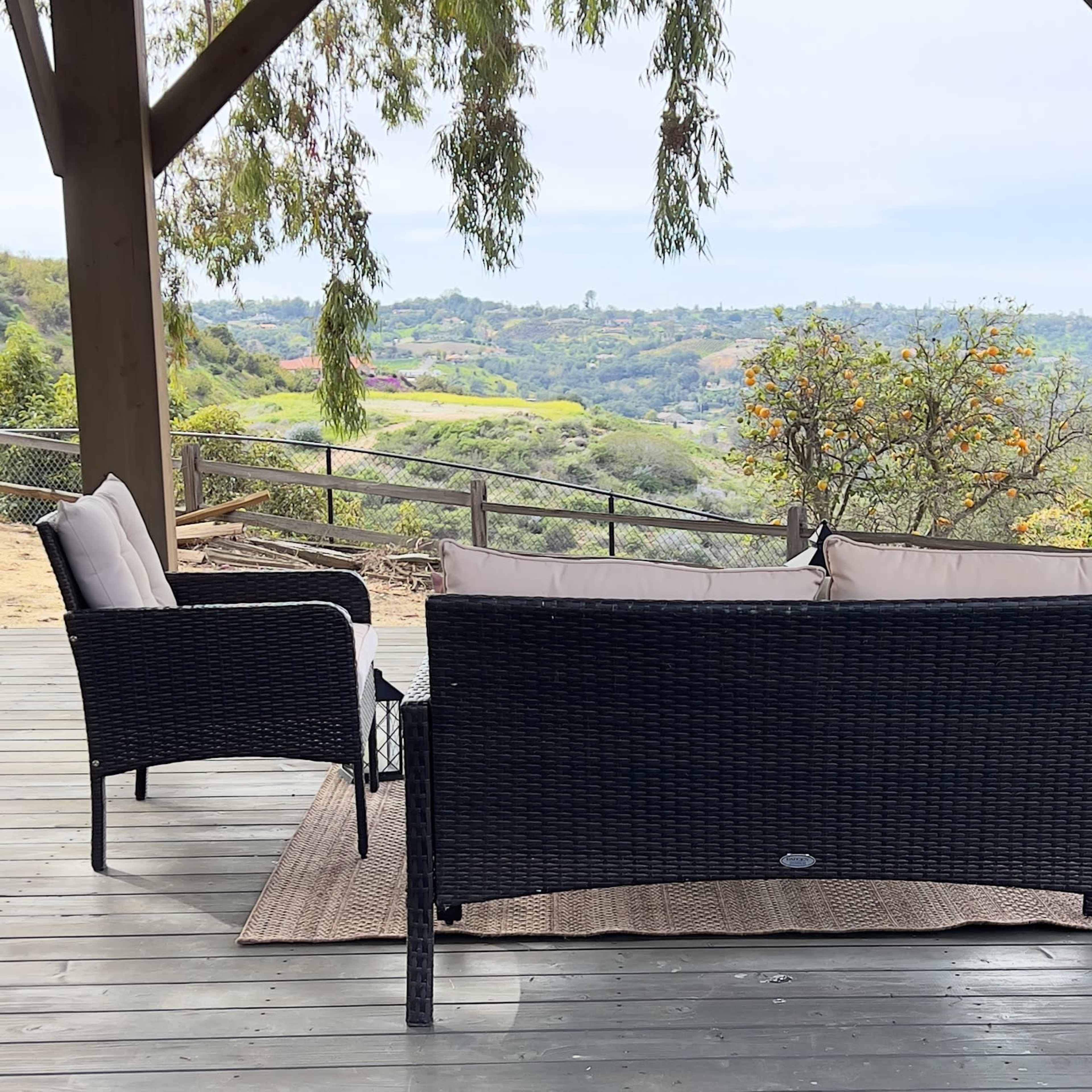 A shaded outdoor seating area features a black wicker sofa and chairs arranged on a wooden deck overlooking a green landscape.