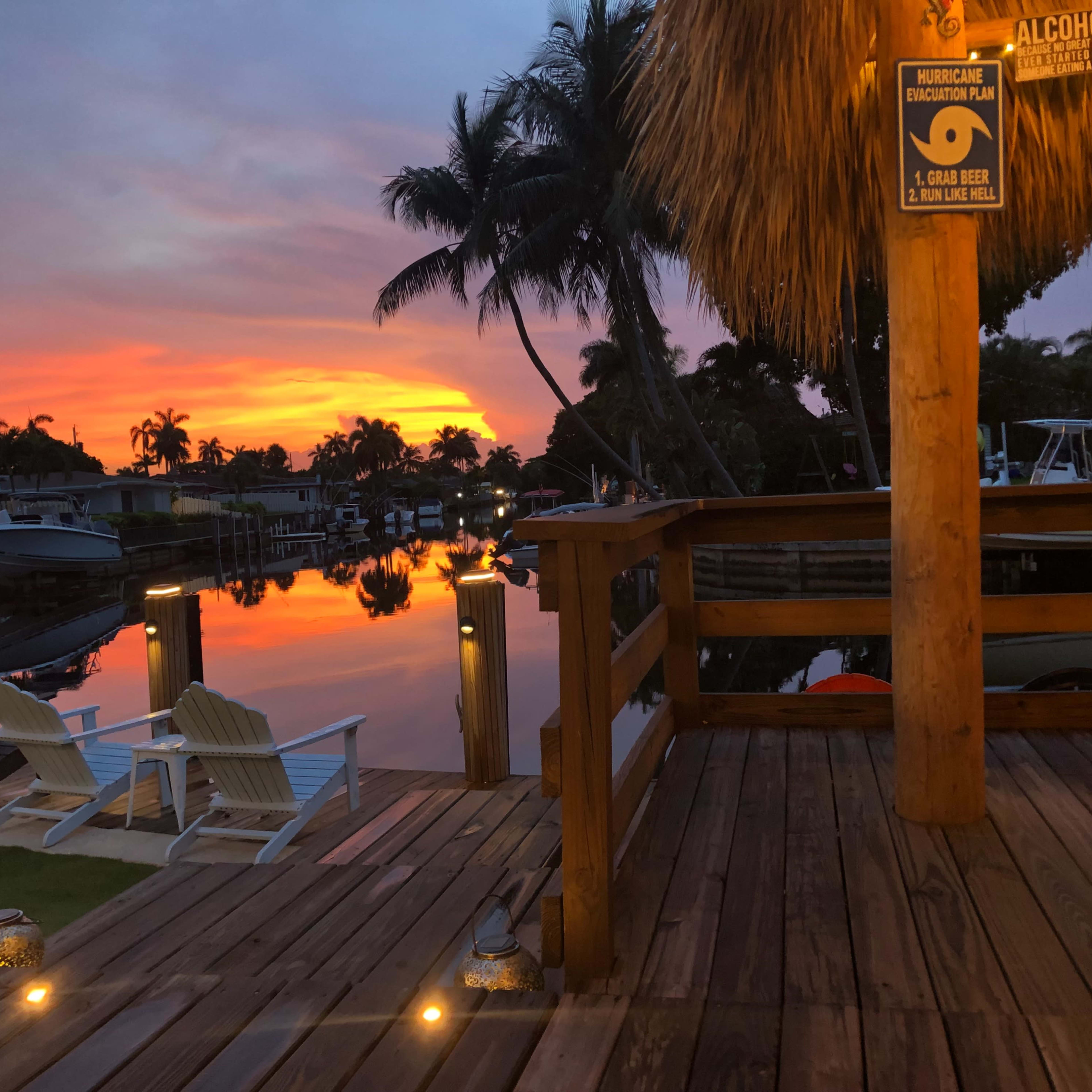 A wooden deck overlooks a calm waterway reflecting a vibrant sunset, with boats docked alongside and palm trees in the background.