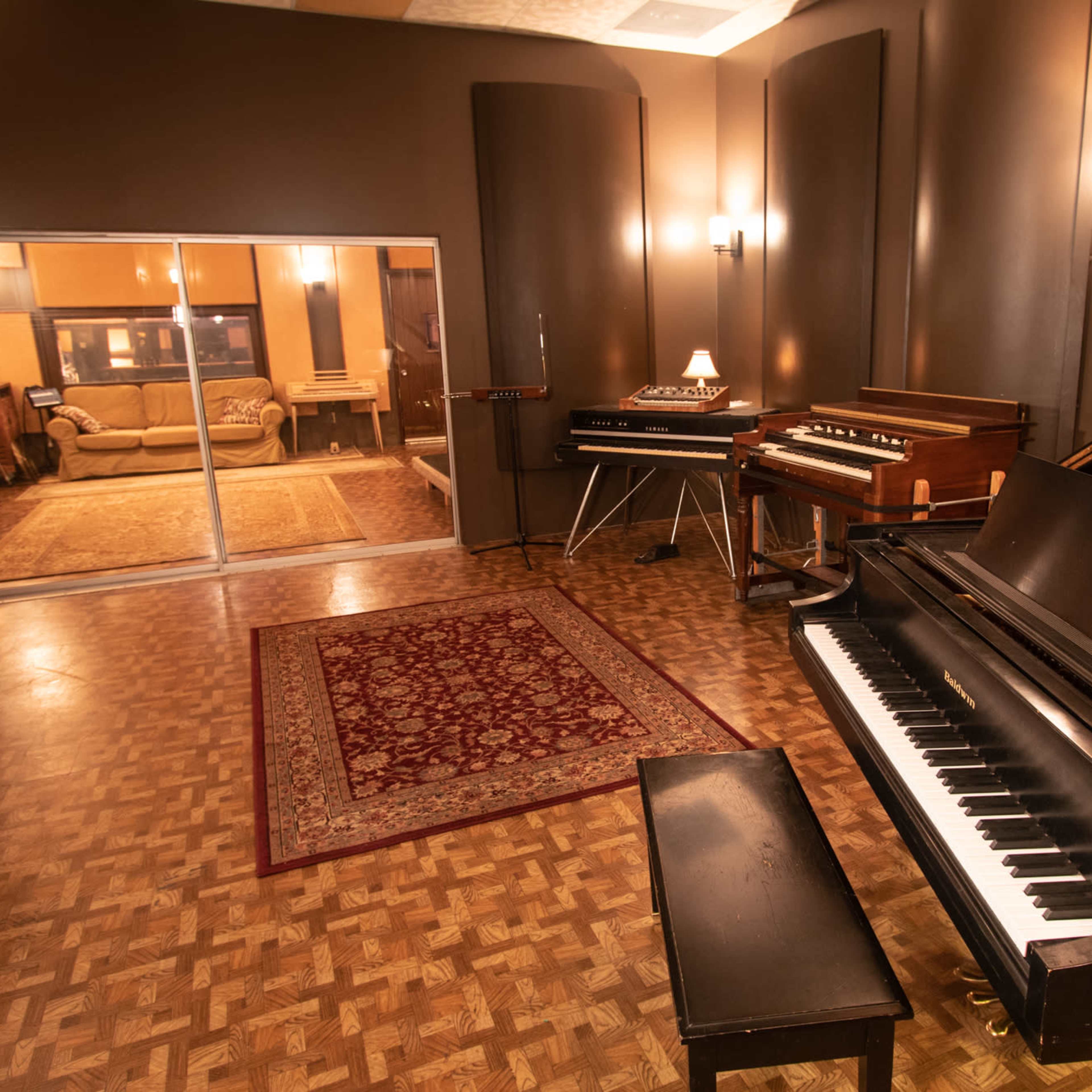 A music studio featuring a grand piano, electronic keyboards, and a cozy seating area visible through large glass doors.