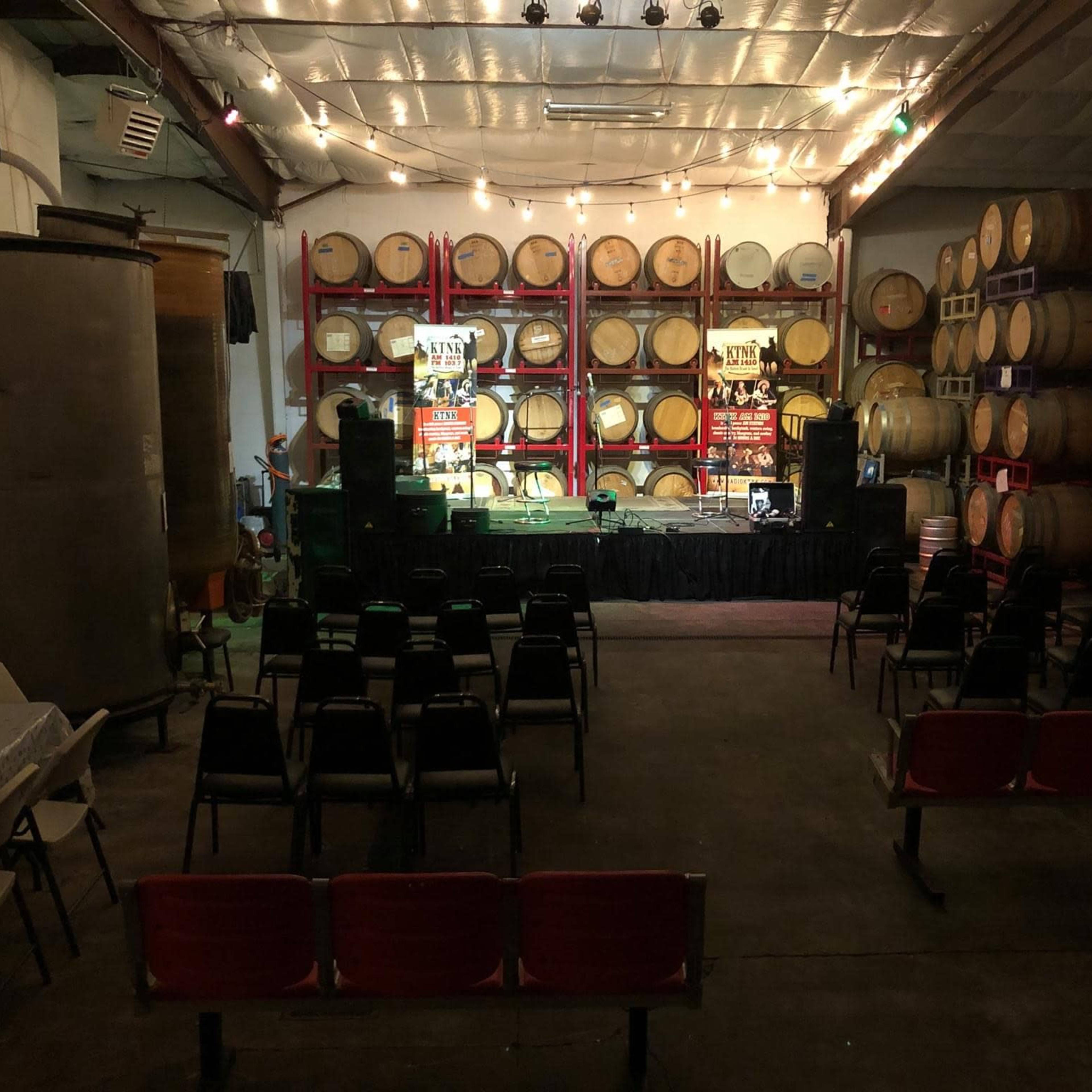 An event space featuring rows of chairs facing a stage, set against a backdrop of stacked wine barrels and ambient lighting.