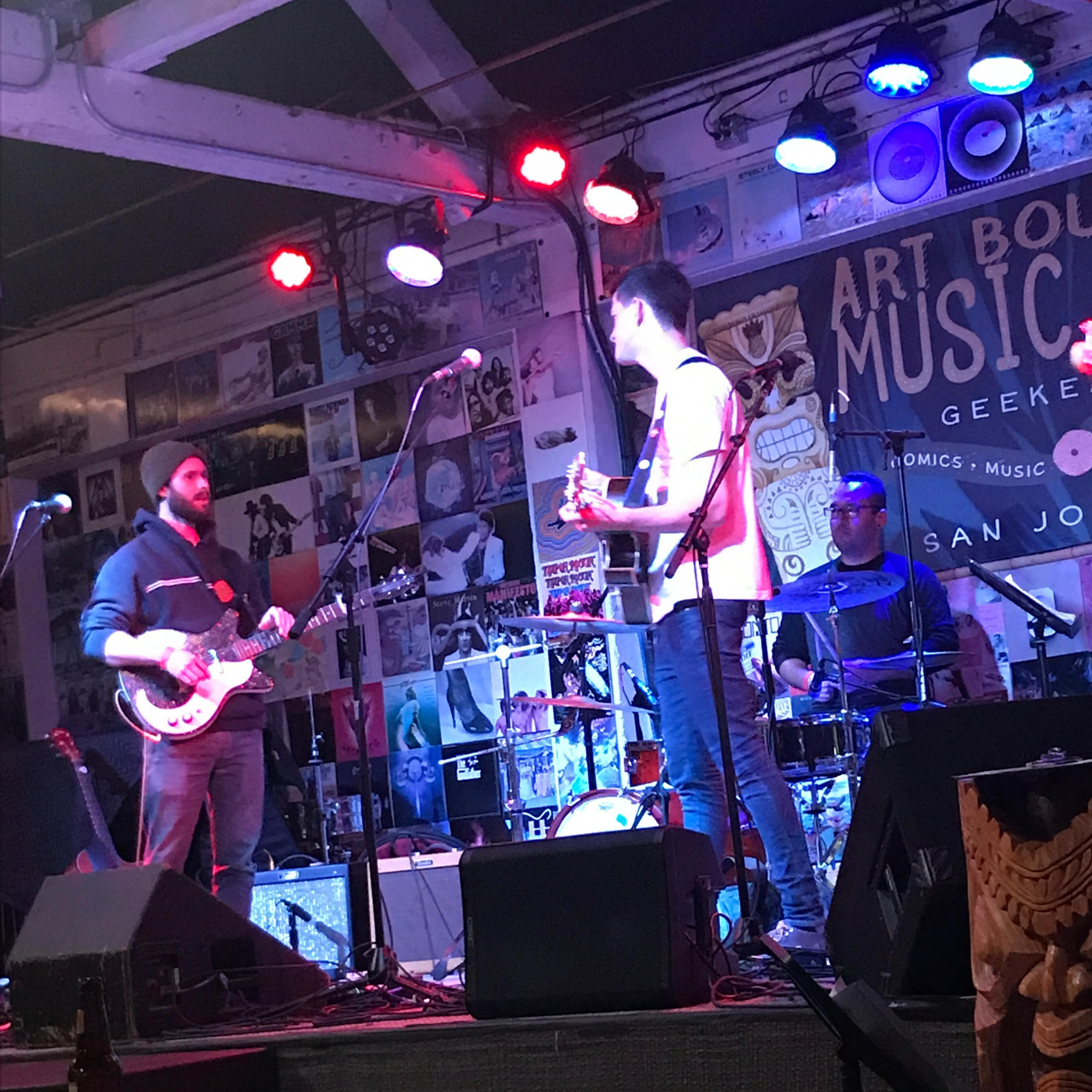 Two musicians perform on stage at a small venue, with colorful lights illuminating the scene and various posters on the walls behind them.