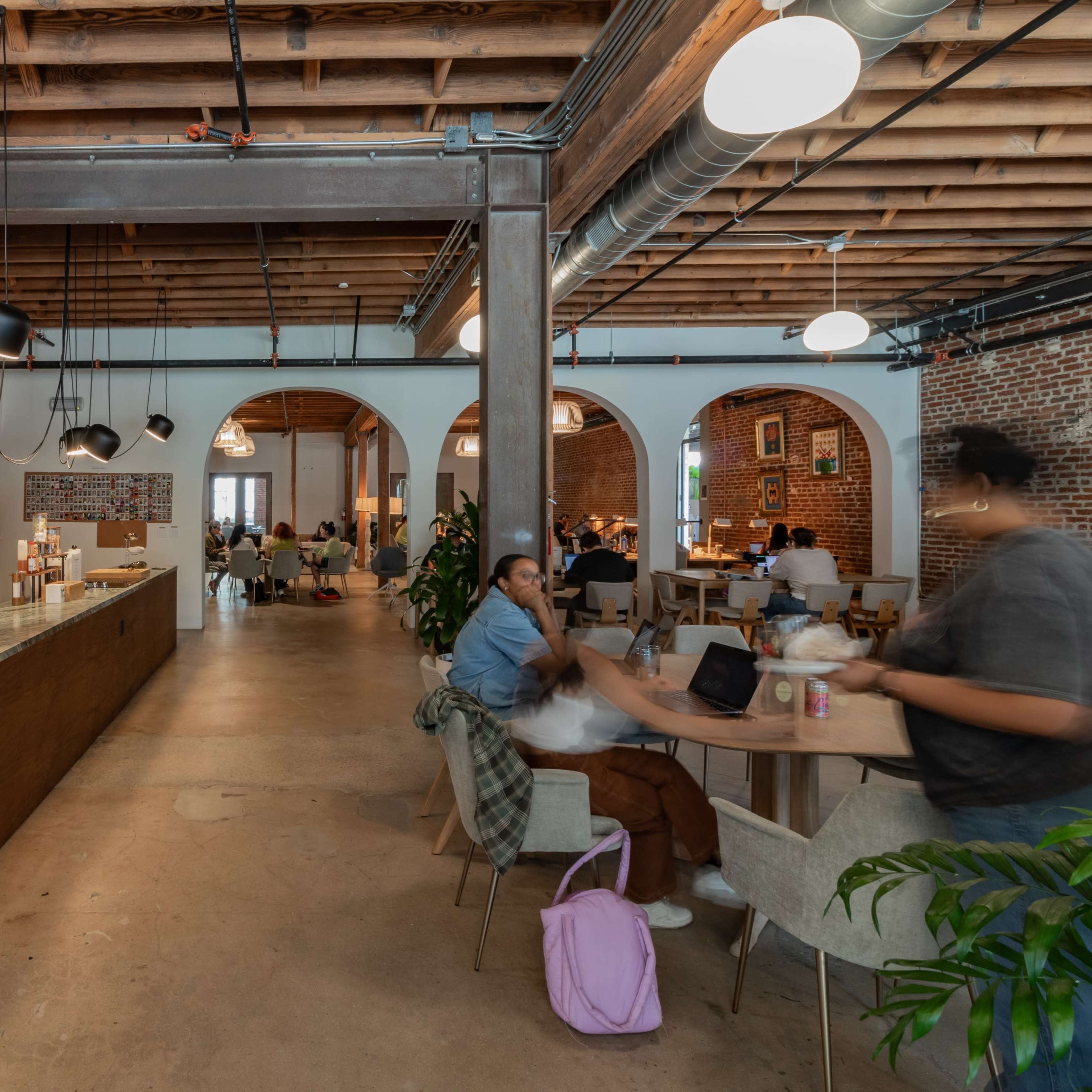 A spacious coworking space with a kitchen area, tables, and people engaged in work and conversations under exposed wooden beams and industrial lighting.