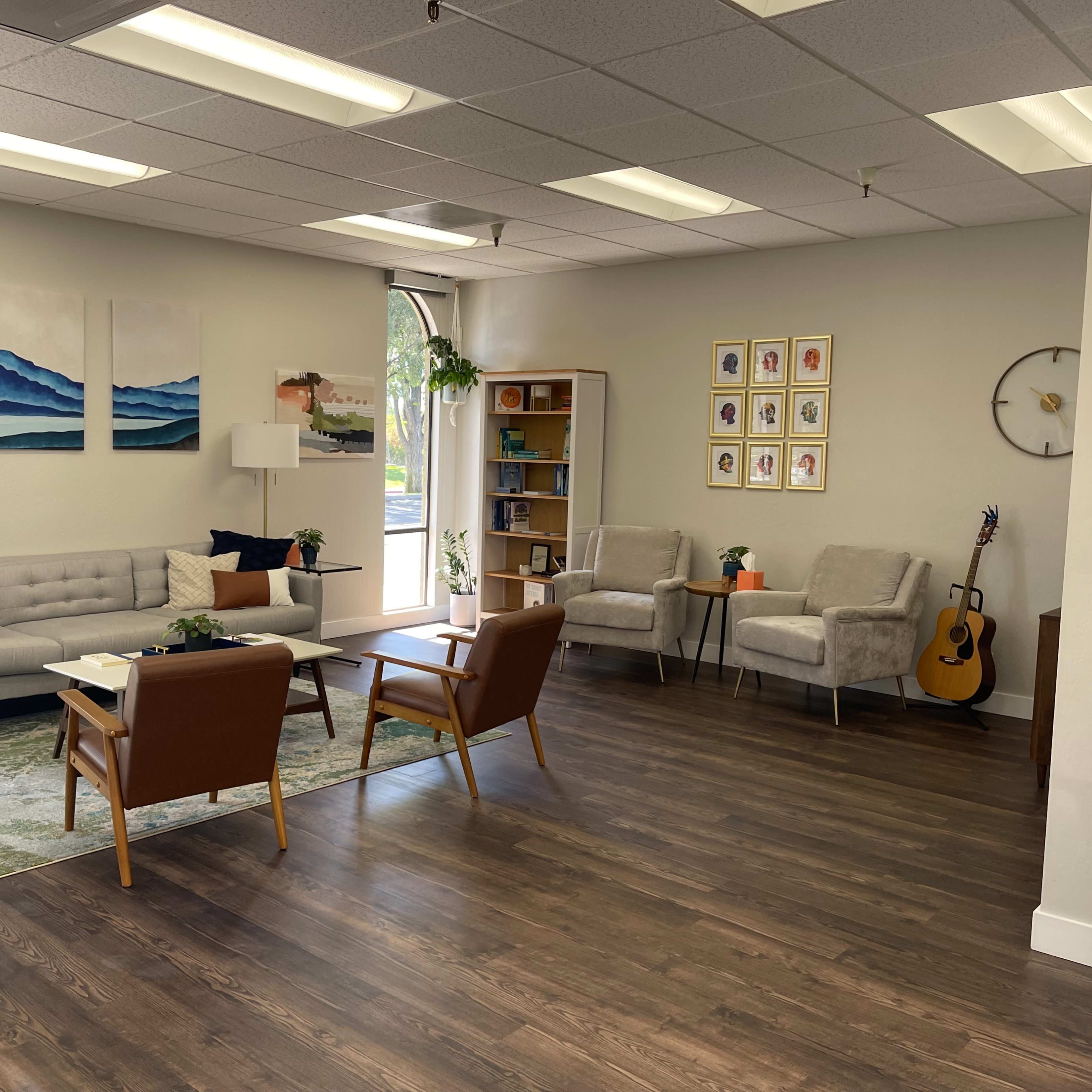 A modern, light-filled waiting area with a seating arrangement, including a sofa, armchairs, and a coffee table, along with artwork on the walls and a guitar in the corner.