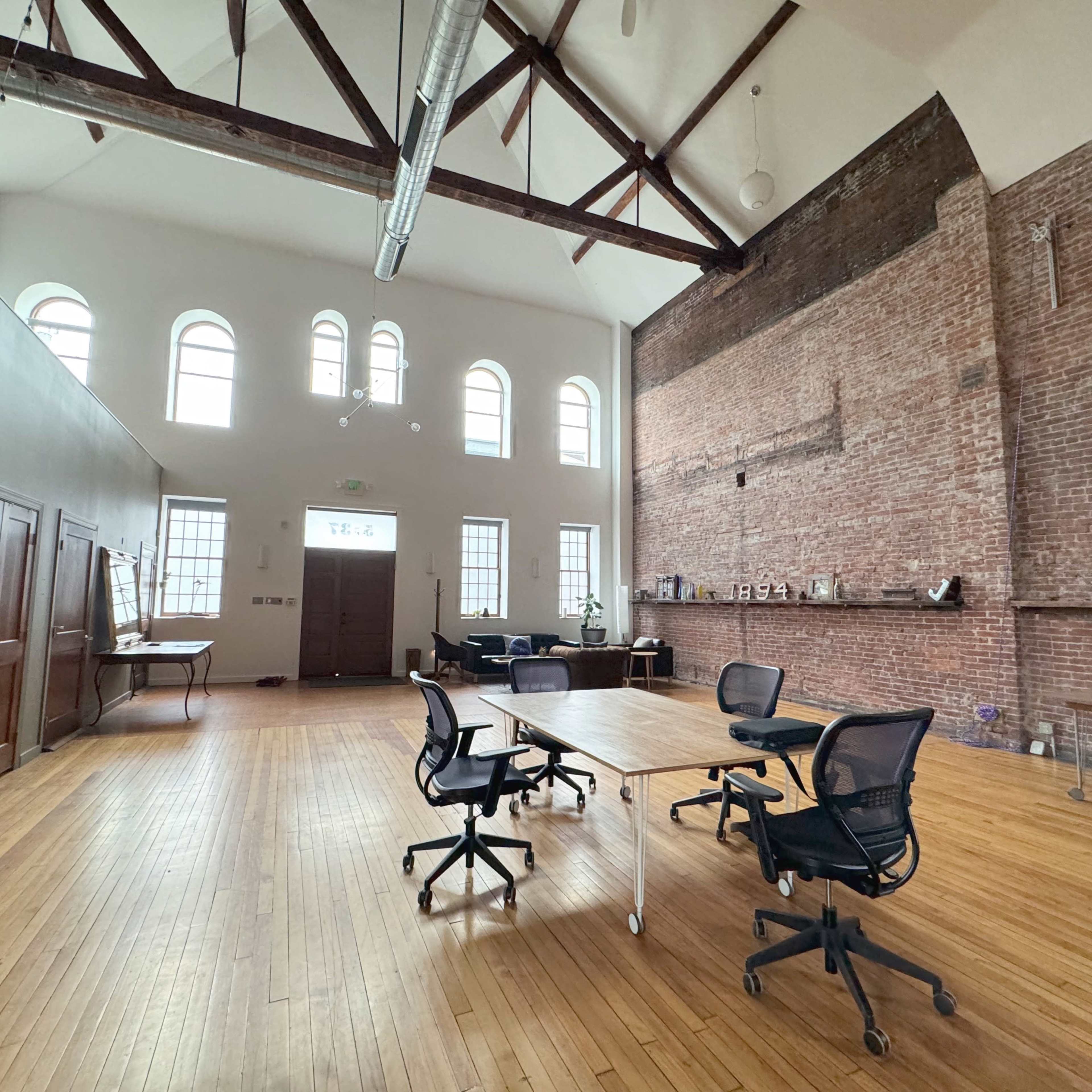 A spacious, modern office with wooden floors, exposed brick walls, and large windows, featuring a table and several rolling chairs.