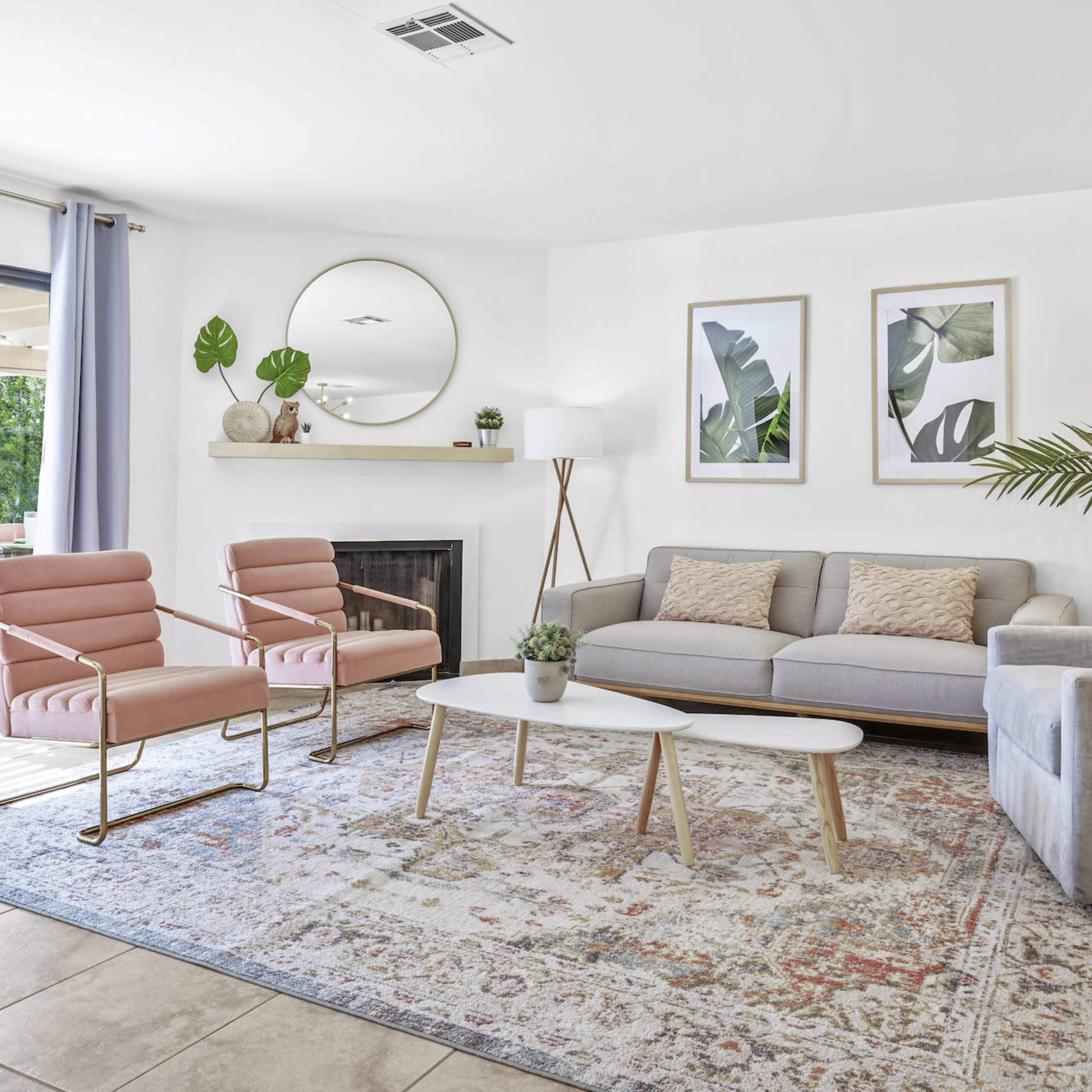A modern living room featuring a gray sofa, two pink accent chairs, a round mirror, and tropical-themed artwork on the walls, with a sliding door leading to a patio.