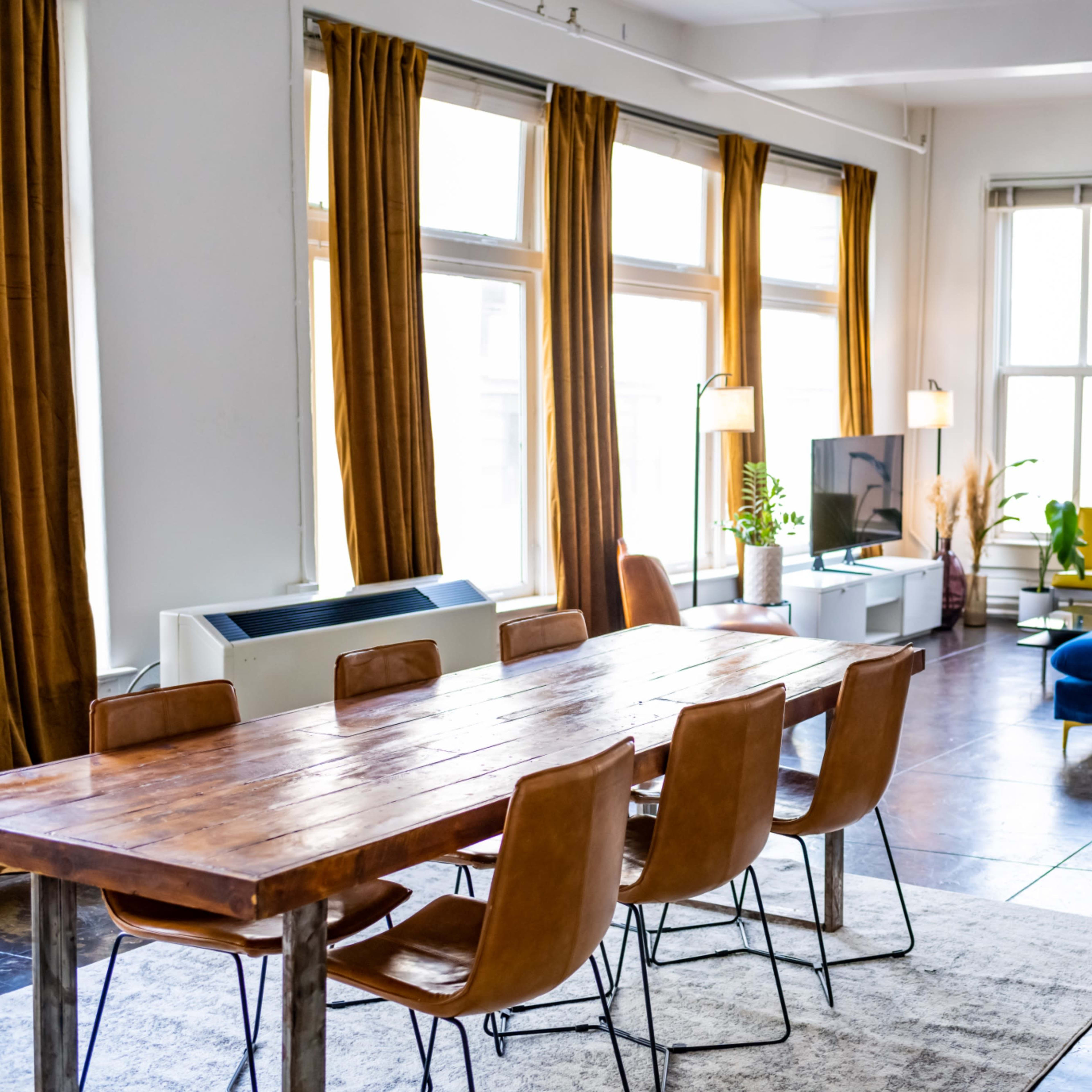 A bright open living space features a wooden dining table with six chairs, a blue sofa, and large windows surrounded by plants.