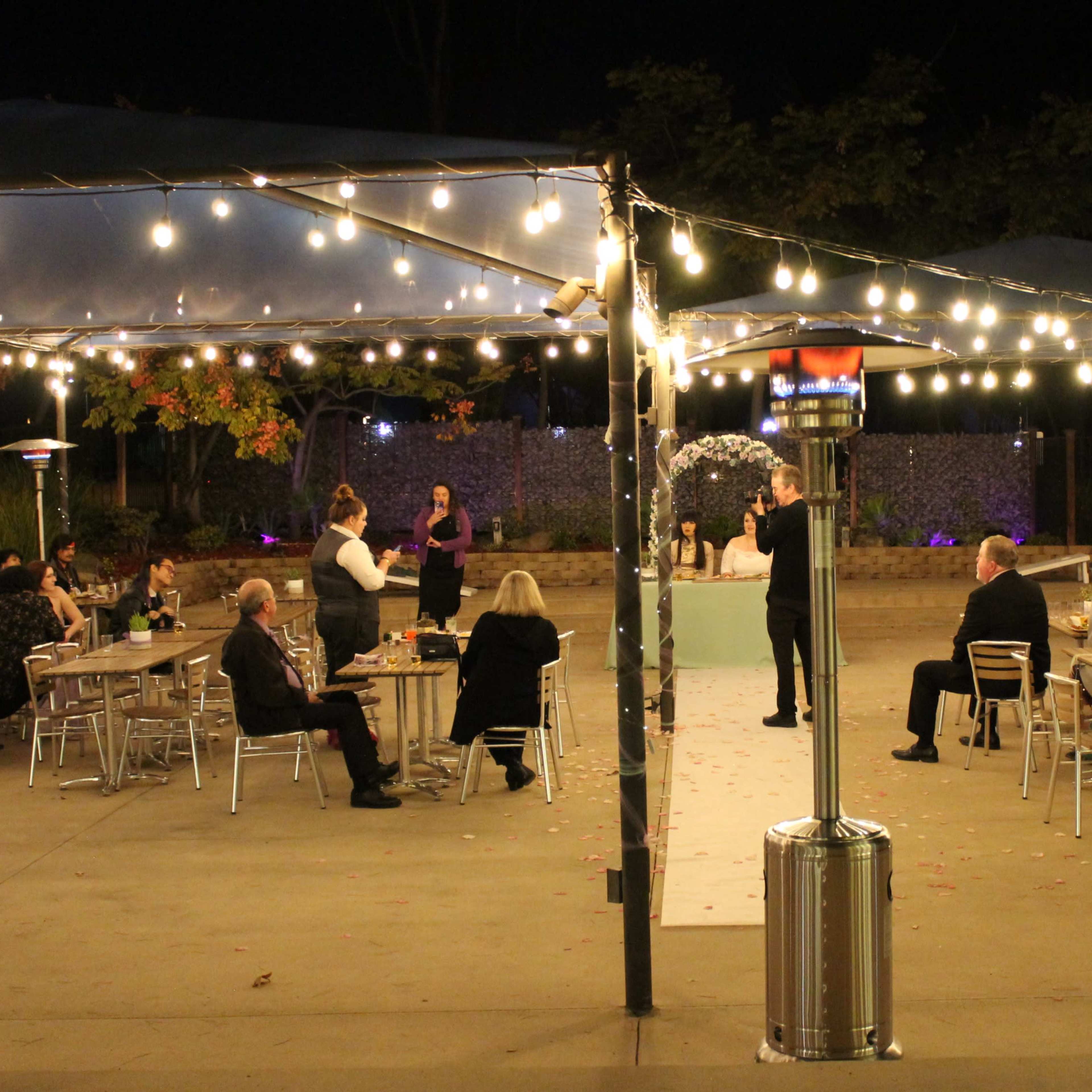A spacious outdoor area is illuminated by string lights, featuring tables and guests socializing under a tent at night.