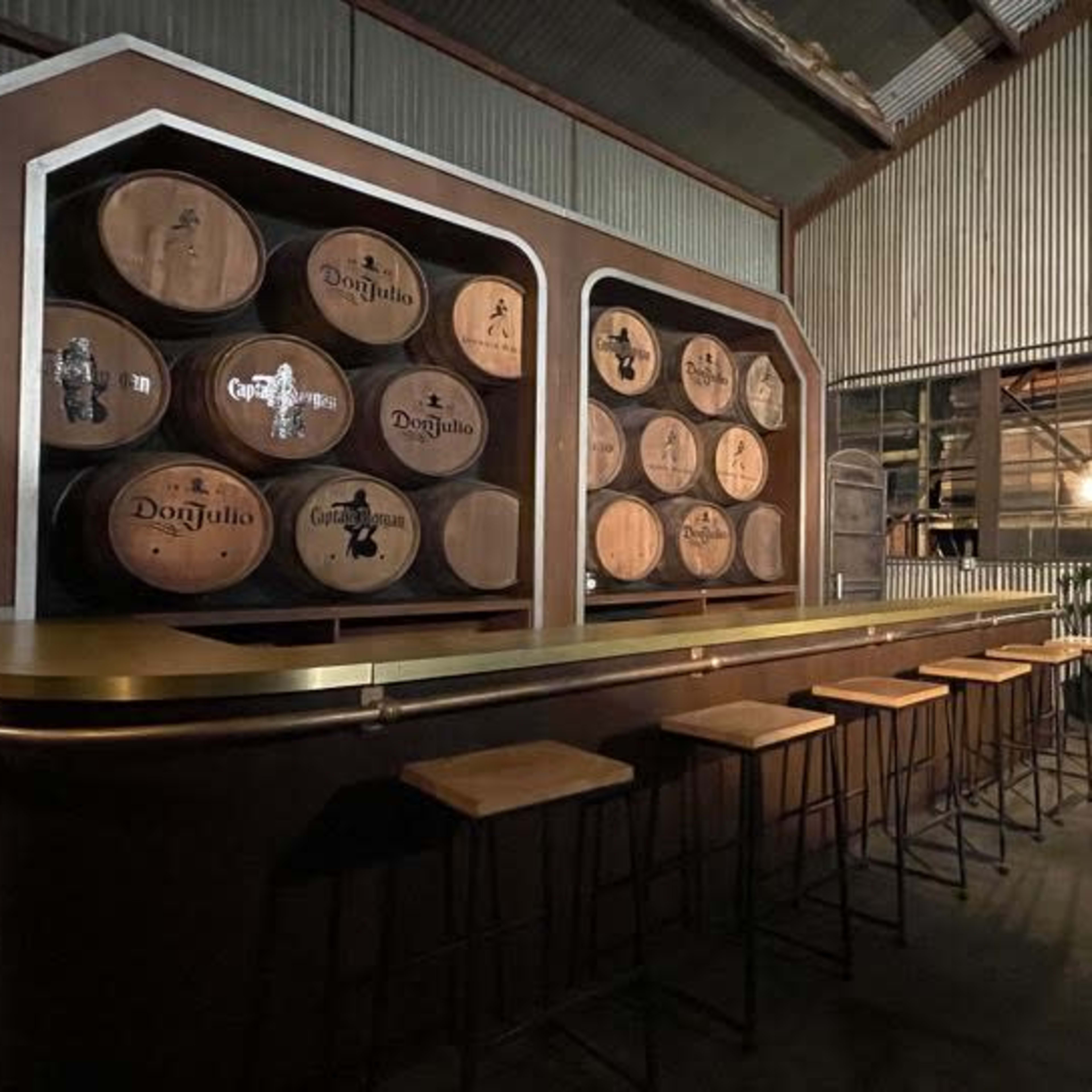A dimly lit bar area with a large wooden counter, several high stools, and a backdrop of wooden barrels mounted on the wall.