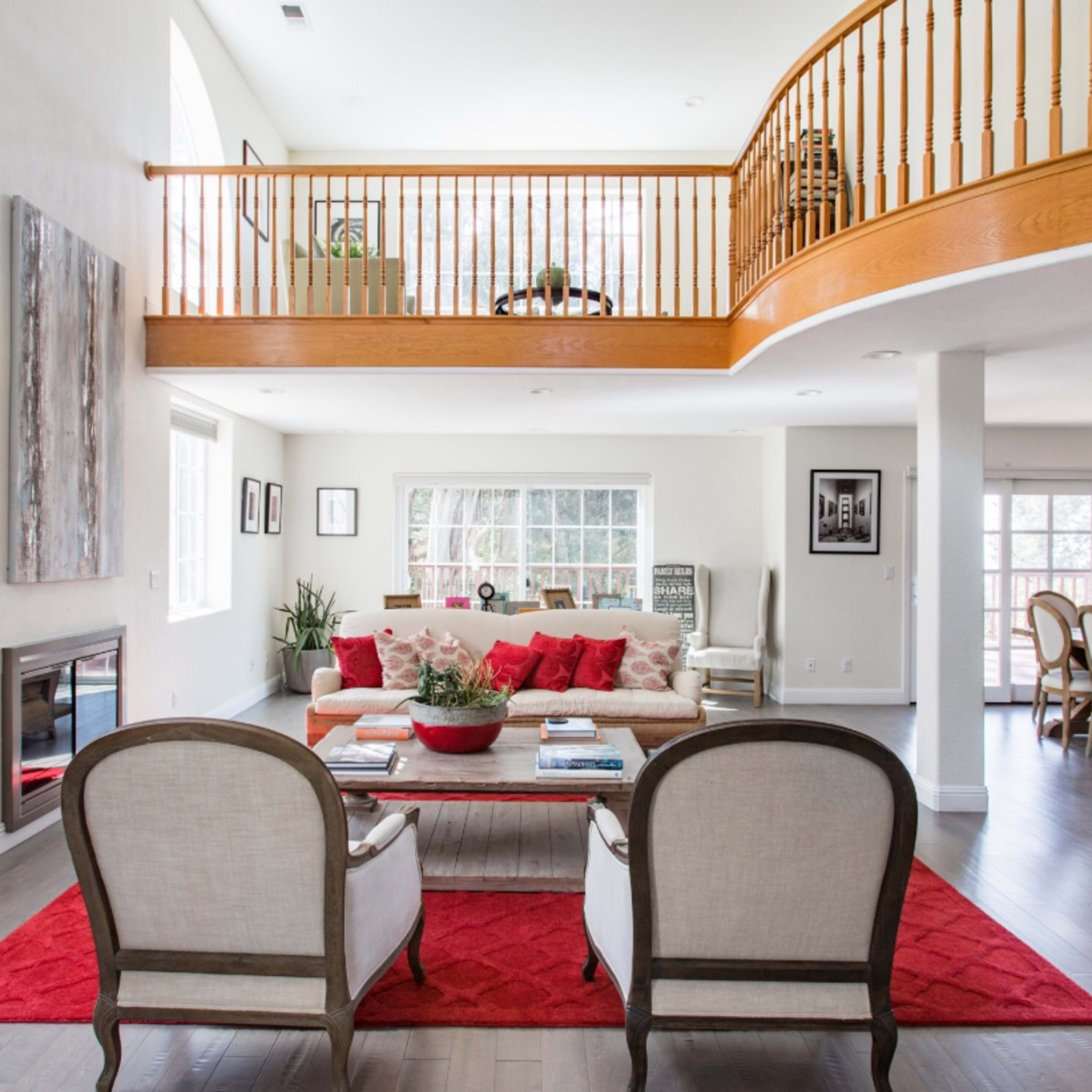 A spacious, well-lit living area featuring a seating arrangement with two chairs, a sofa adorned with red cushions, and an upper balcony overlooking the space.