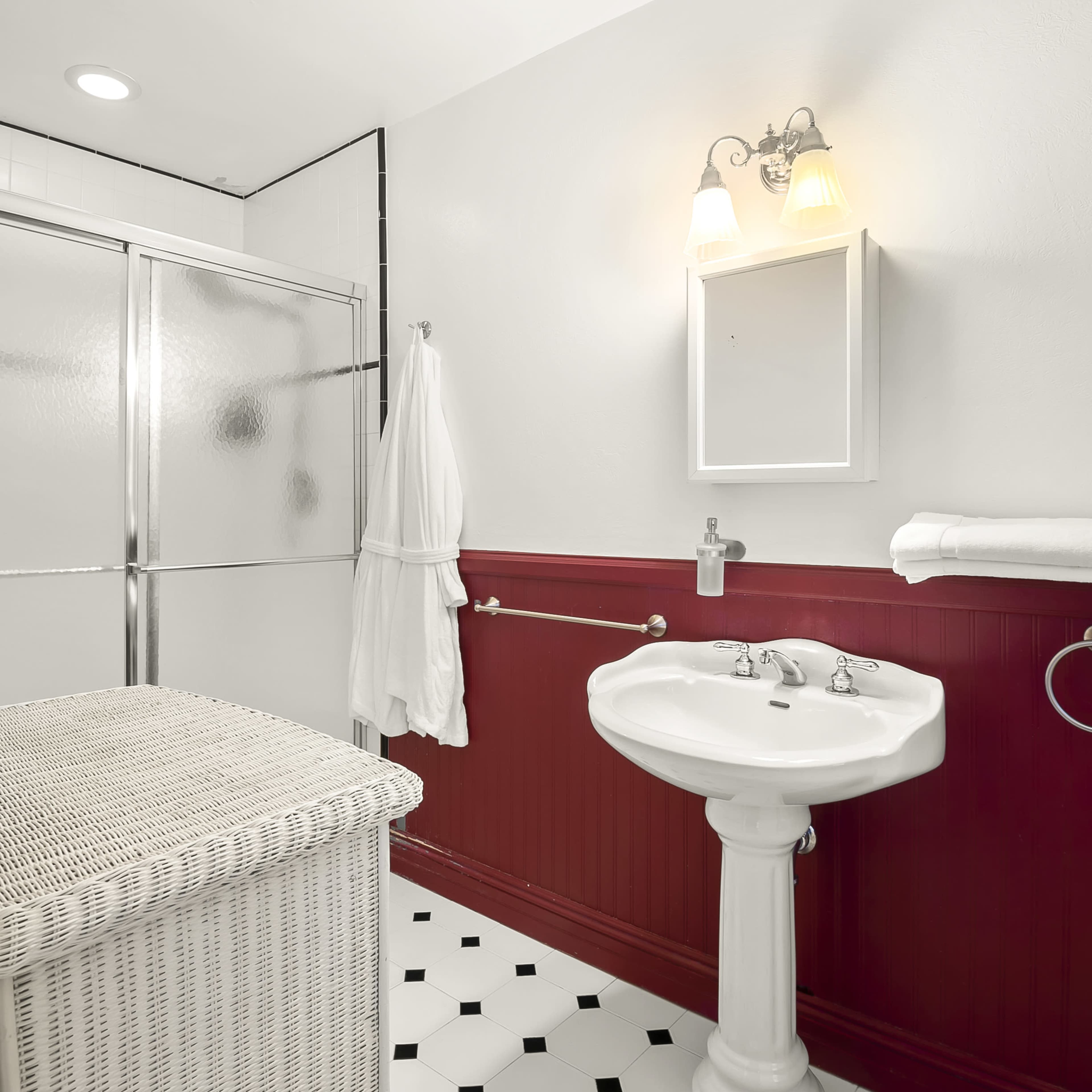 A bathroom with a shower area, a white pedestal sink, a wicker laundry basket, and a light fixture above a mirror, all featuring a red and white color scheme.