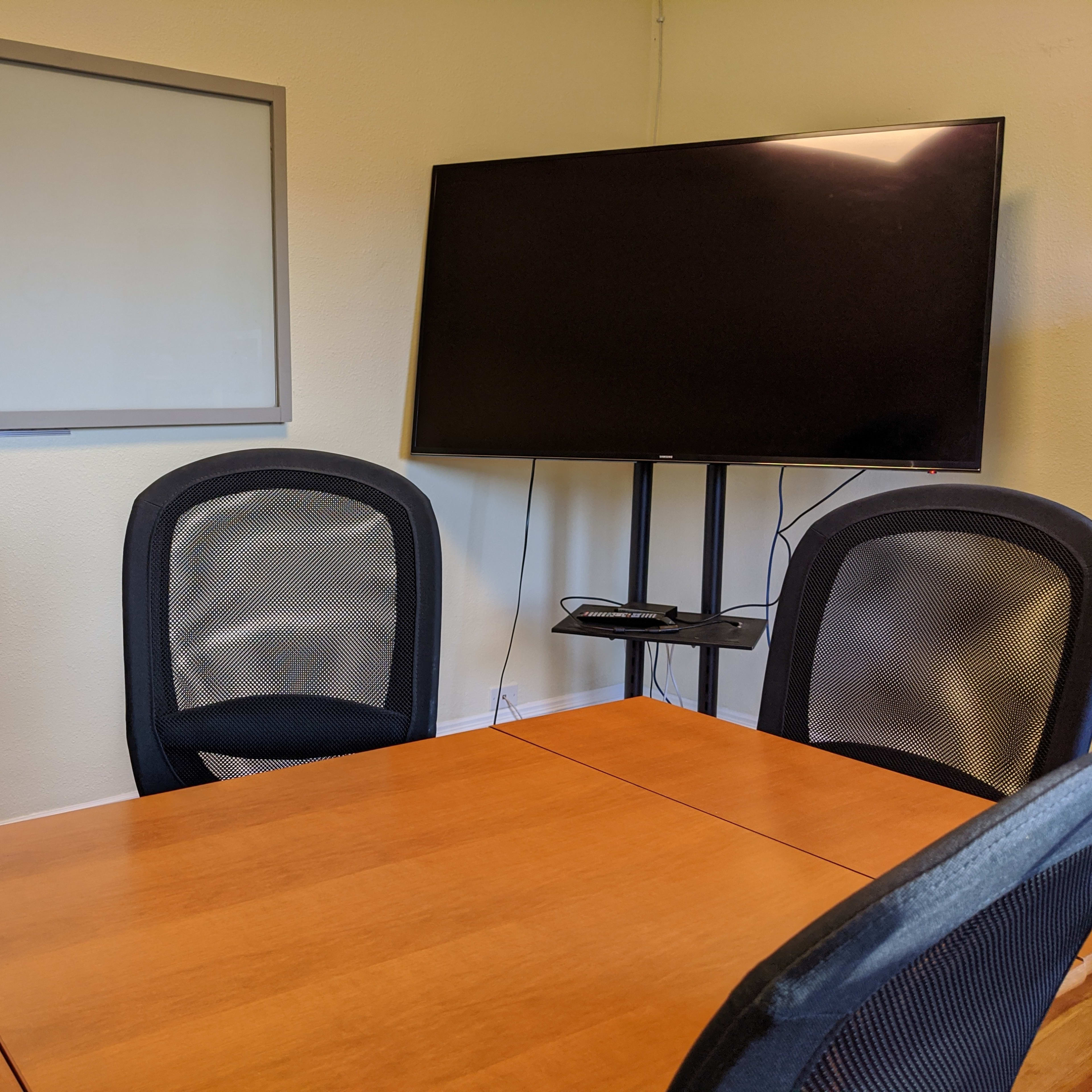 A conference room with a brown table, four black mesh chairs, a whiteboard, and a wall-mounted television.
