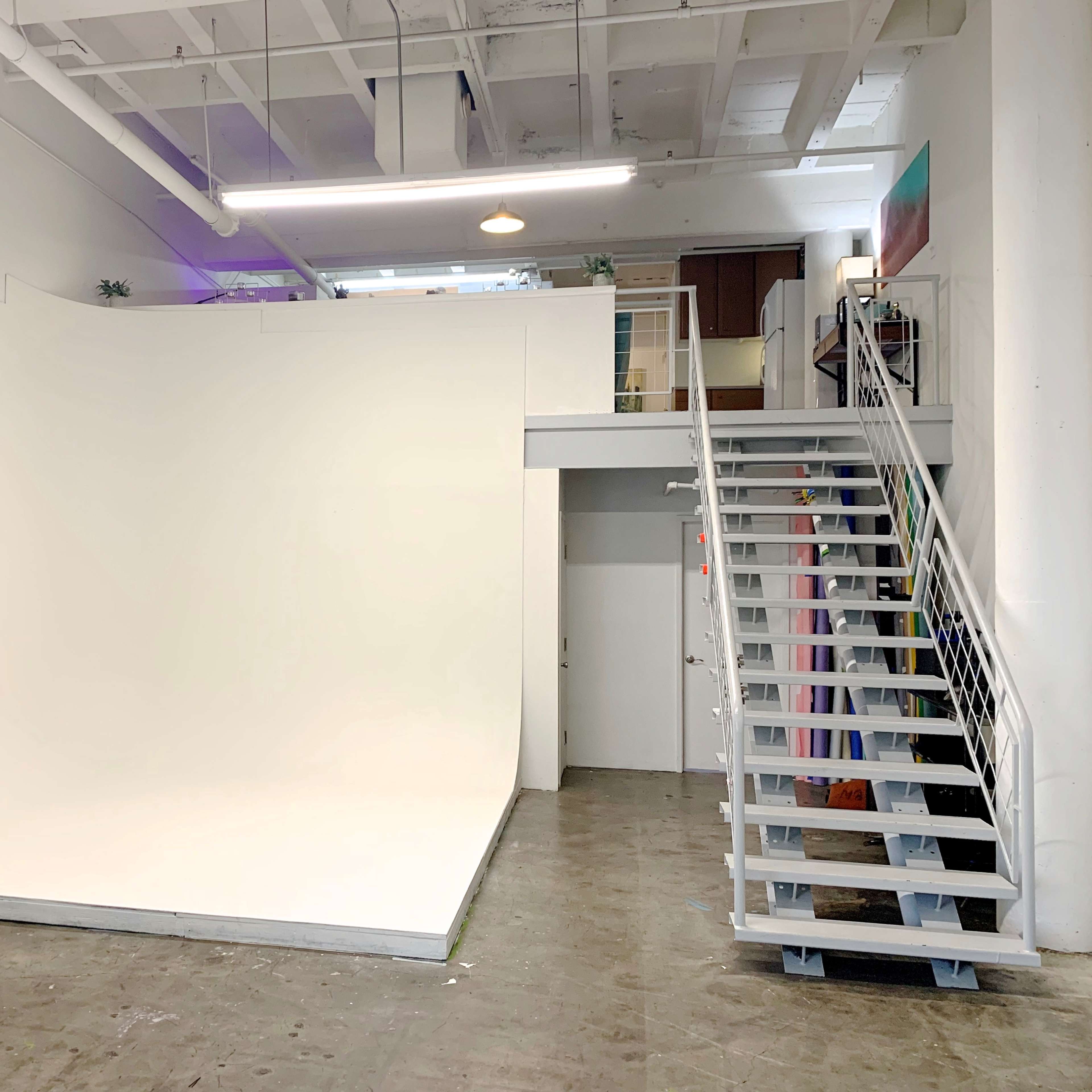 A bright, minimalist studio space with a large white background and a staircase leading to an upper level.