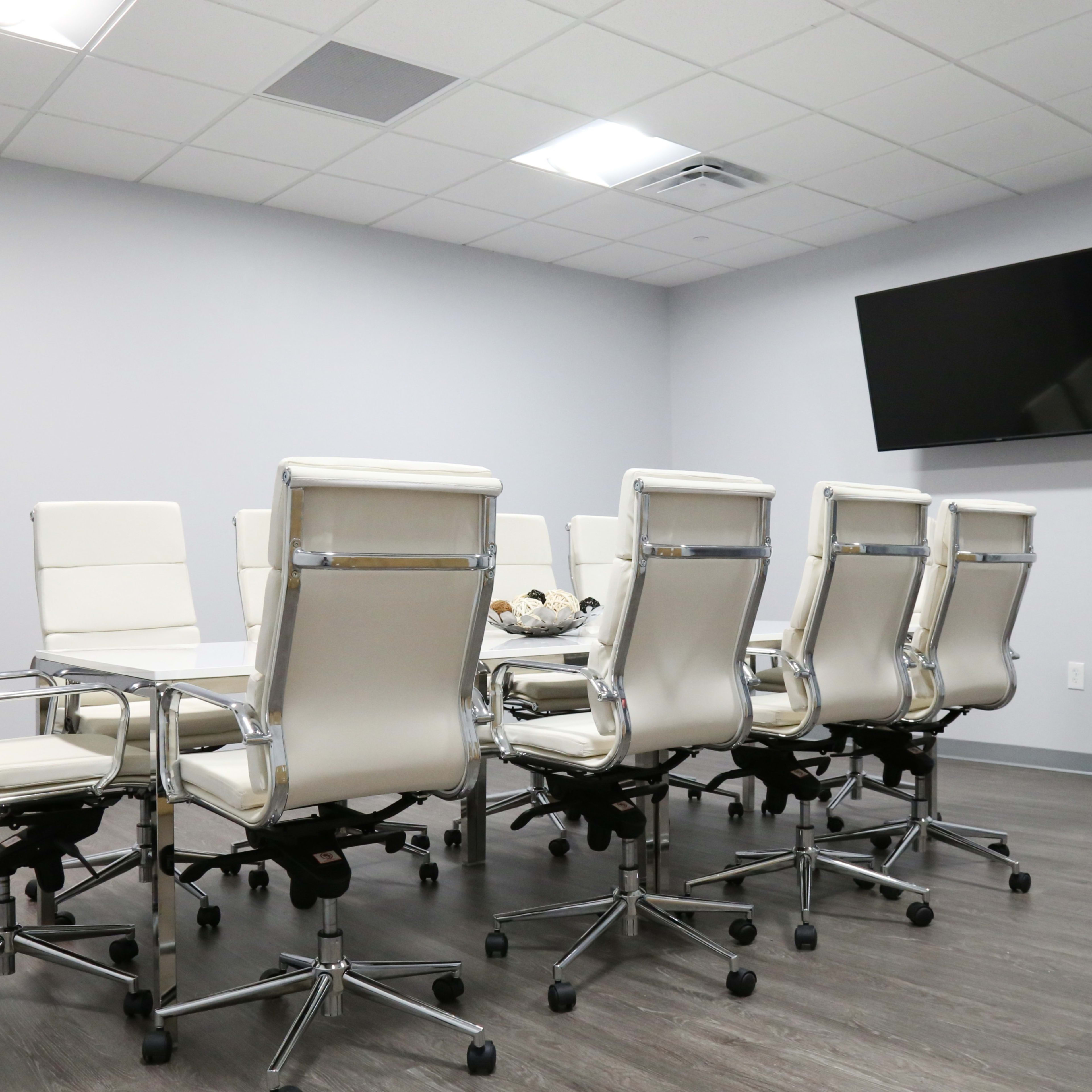 A modern conference room features a long table surrounded by eight white leather office chairs and a wall-mounted television.