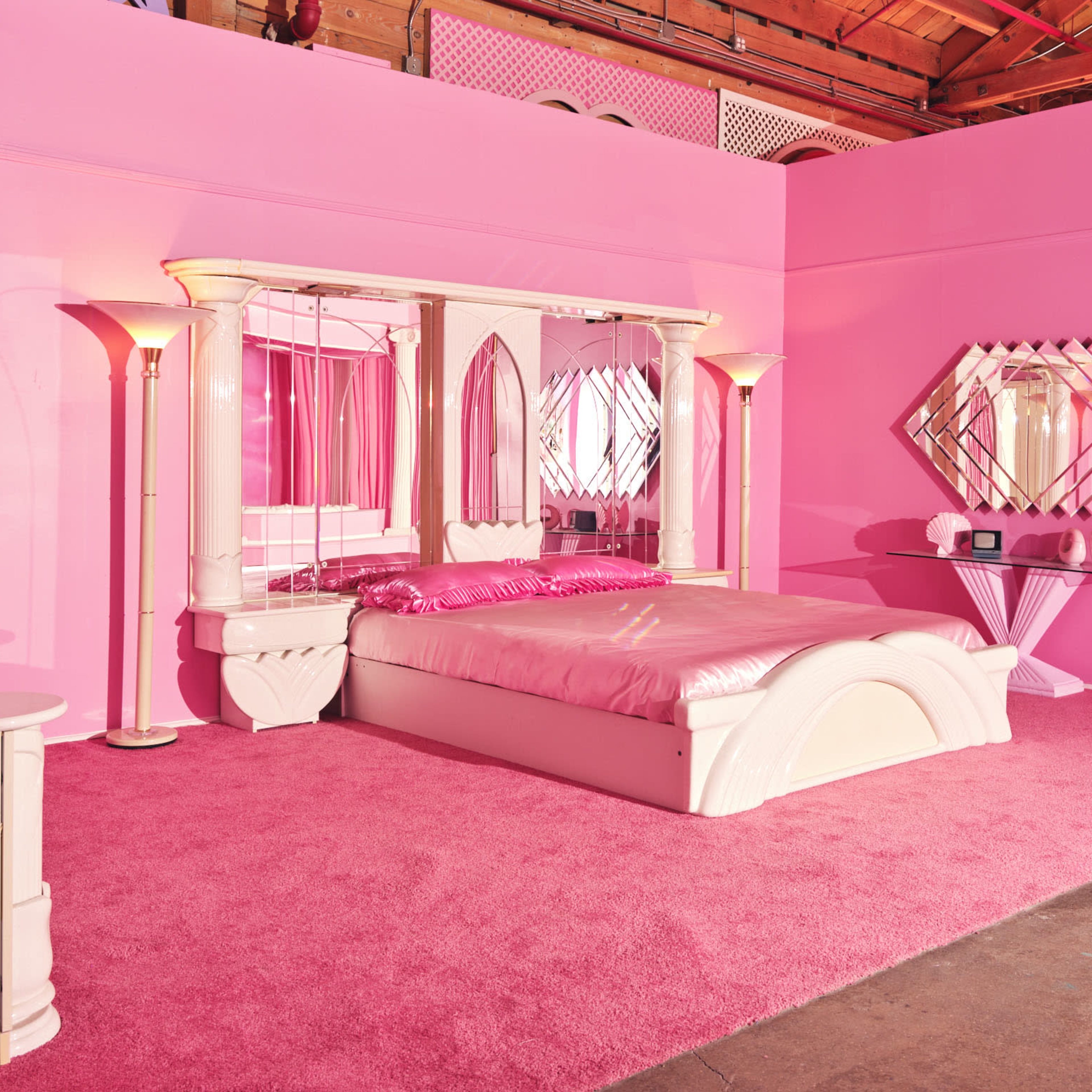 A pink-themed bedroom featuring a bed with a pink comforter, matching walls, and mirrored furniture.