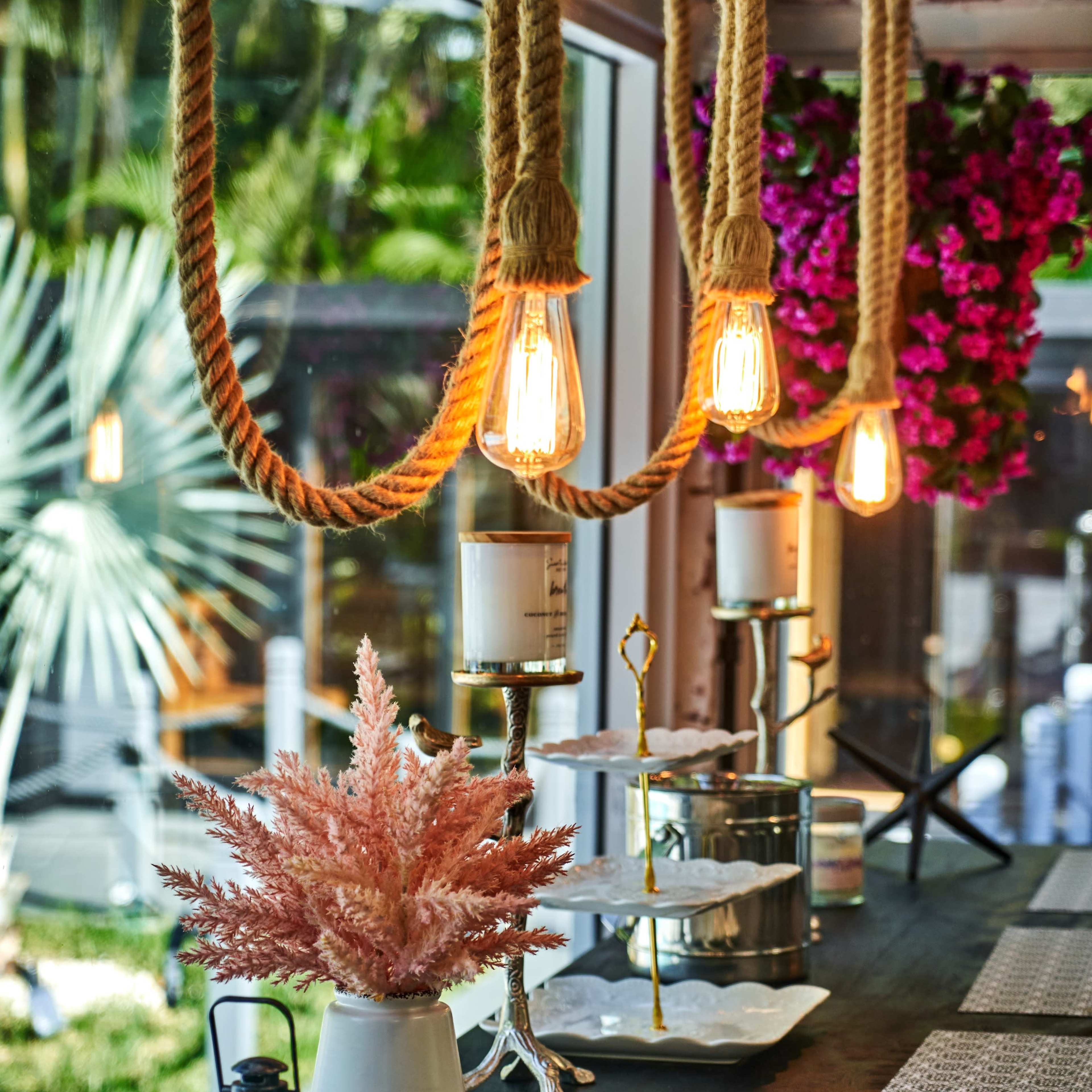 A stylish indoor setting with pendant lights hanging from ropes, a low wooden table adorned with decorative plants, and a background of lush greenery and flowers.