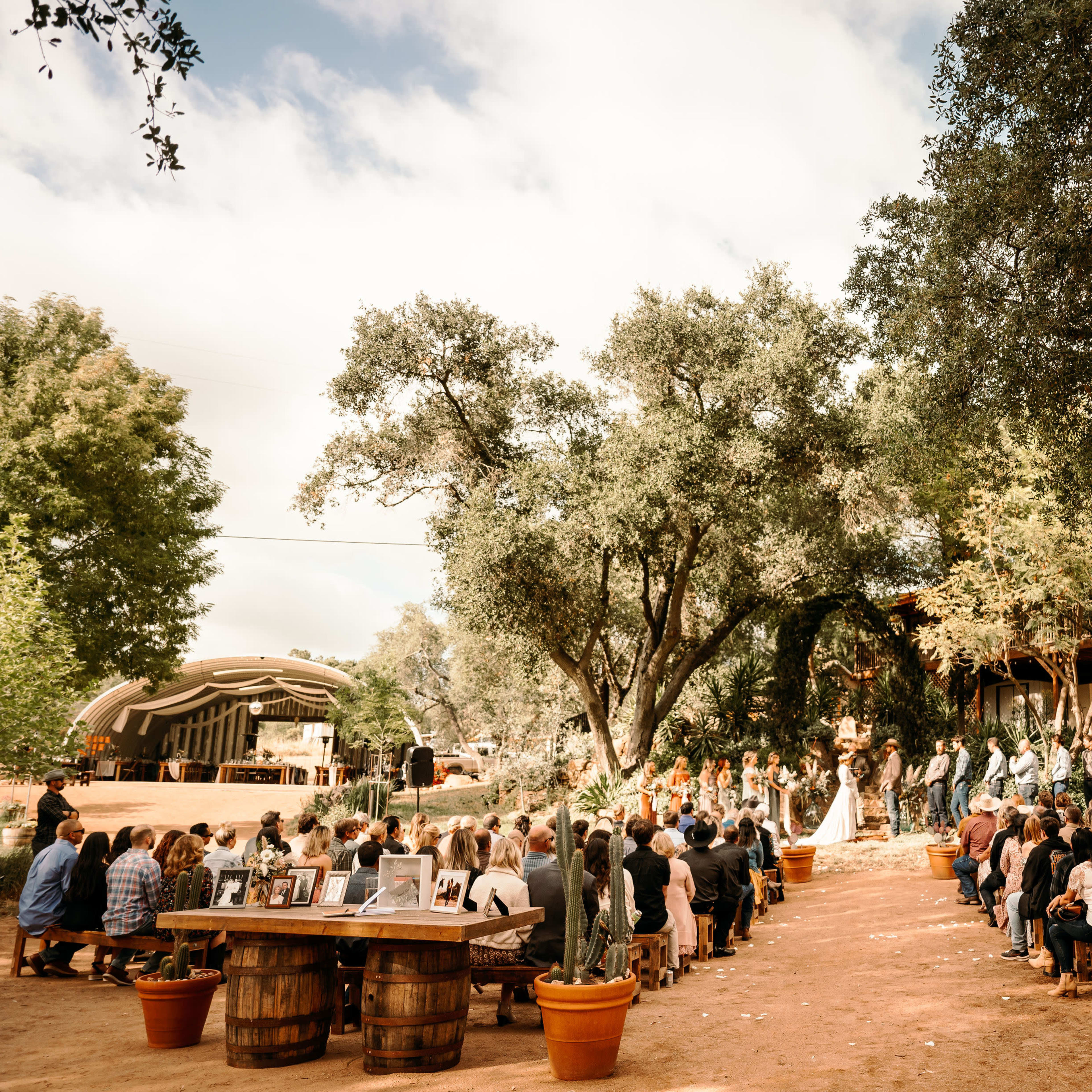 A wedding ceremony takes place outdoors under a large tree, with guests seated on benches facing the altar and a decorated stage in the background.