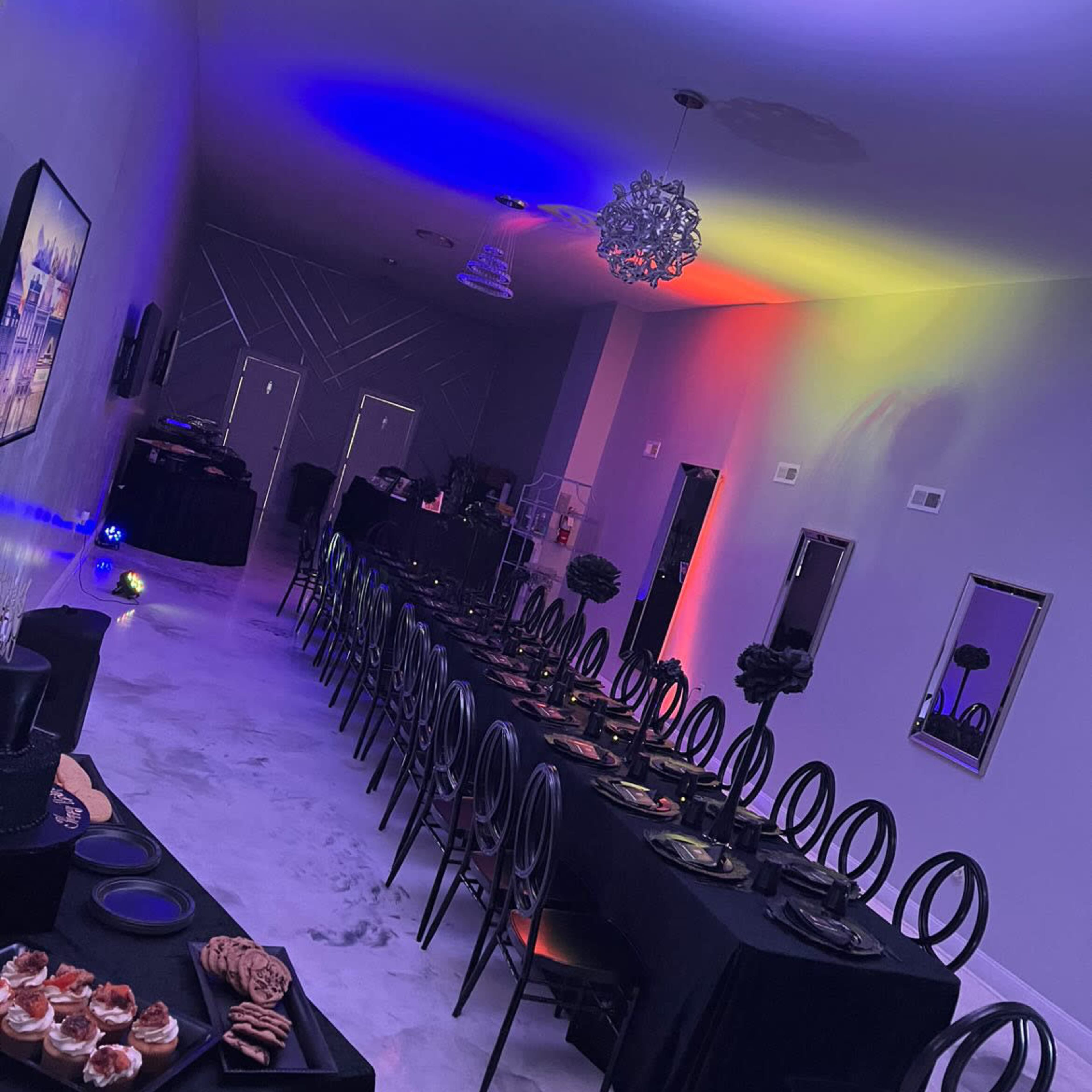 A long, elegantly set dining table is arranged in a decorated room with purple and orange lighting.