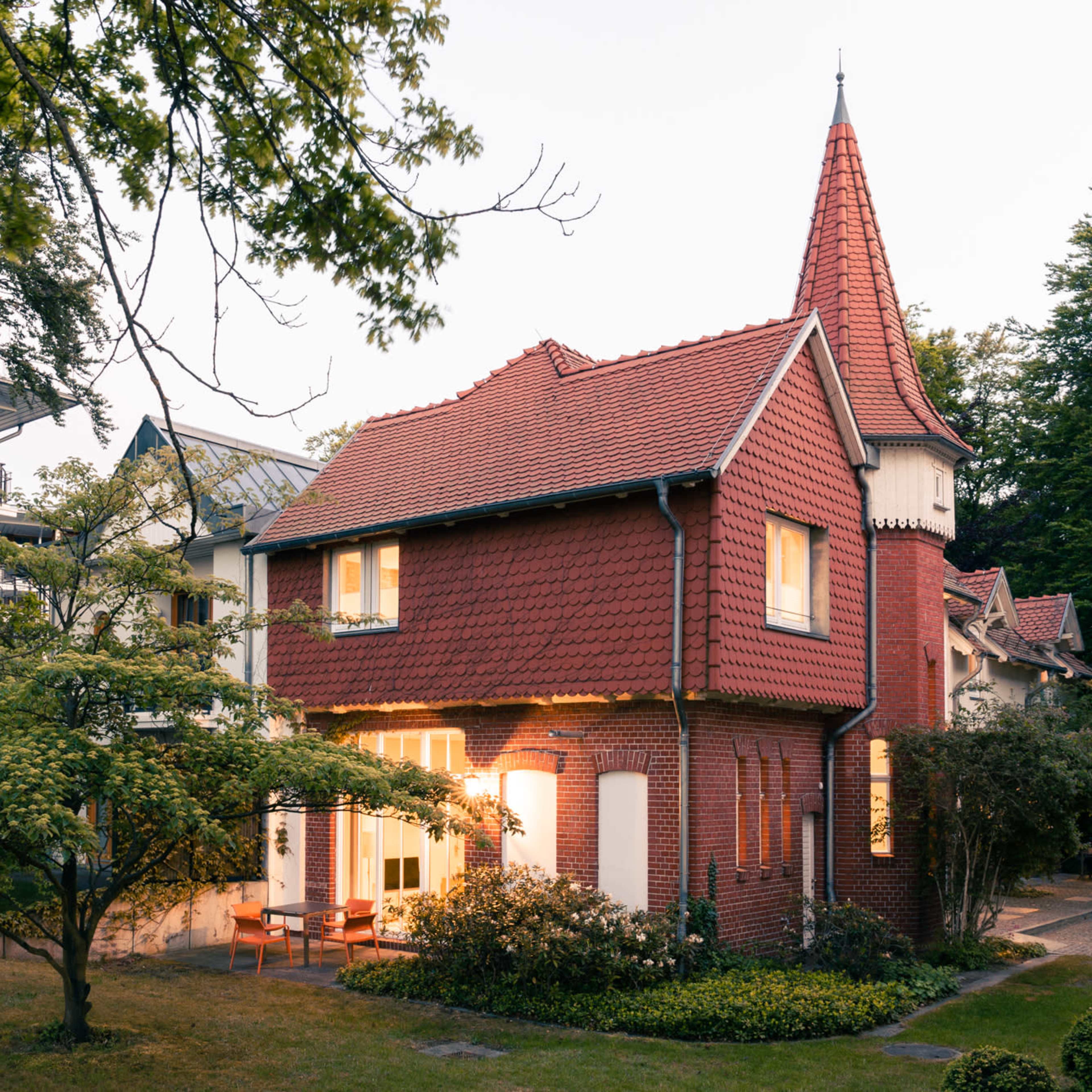 A red-roofed house with a pointed tower is surrounded by trees and greenery.